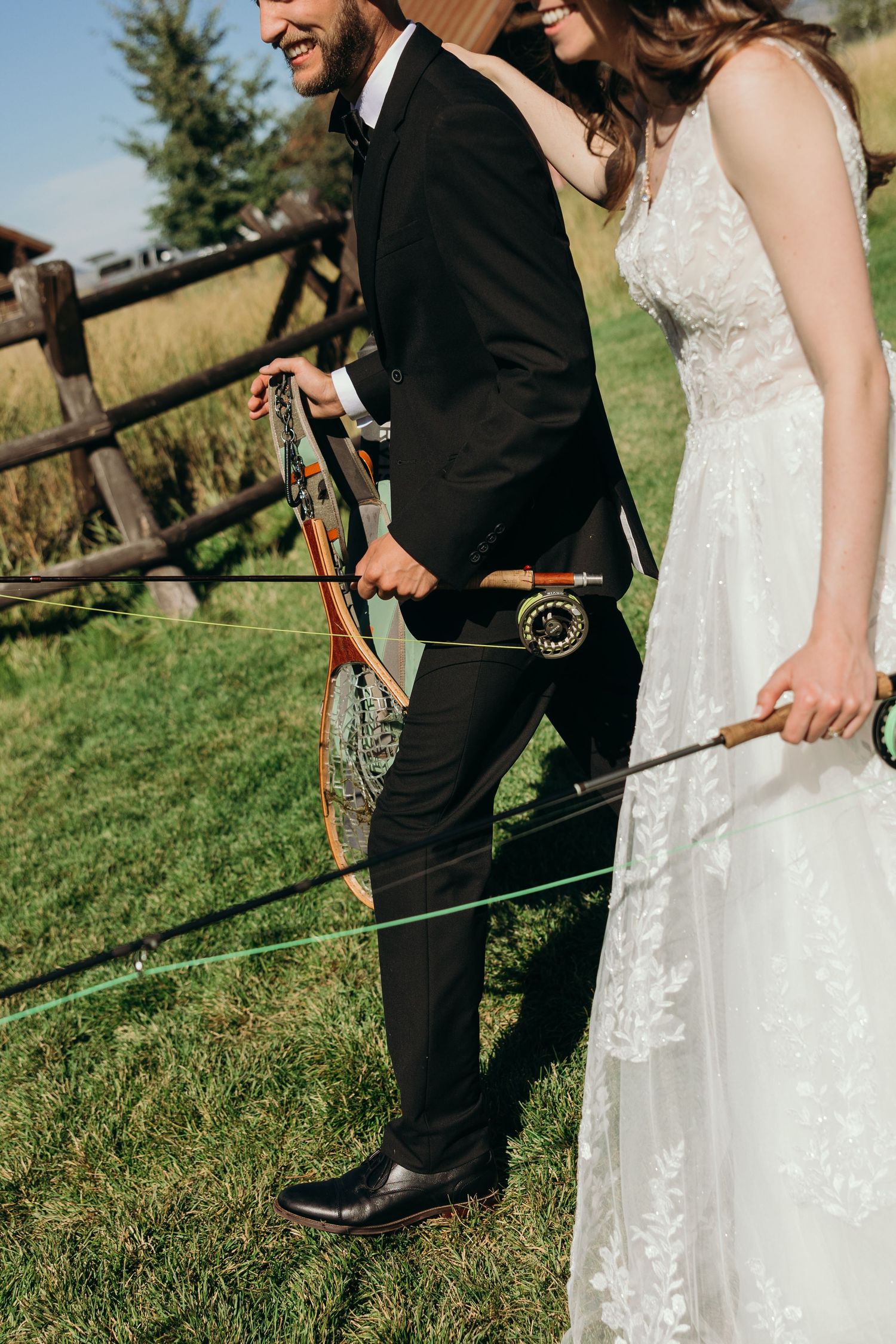 A couple in wedding attire stands by a rustic fence holding archery equipment.