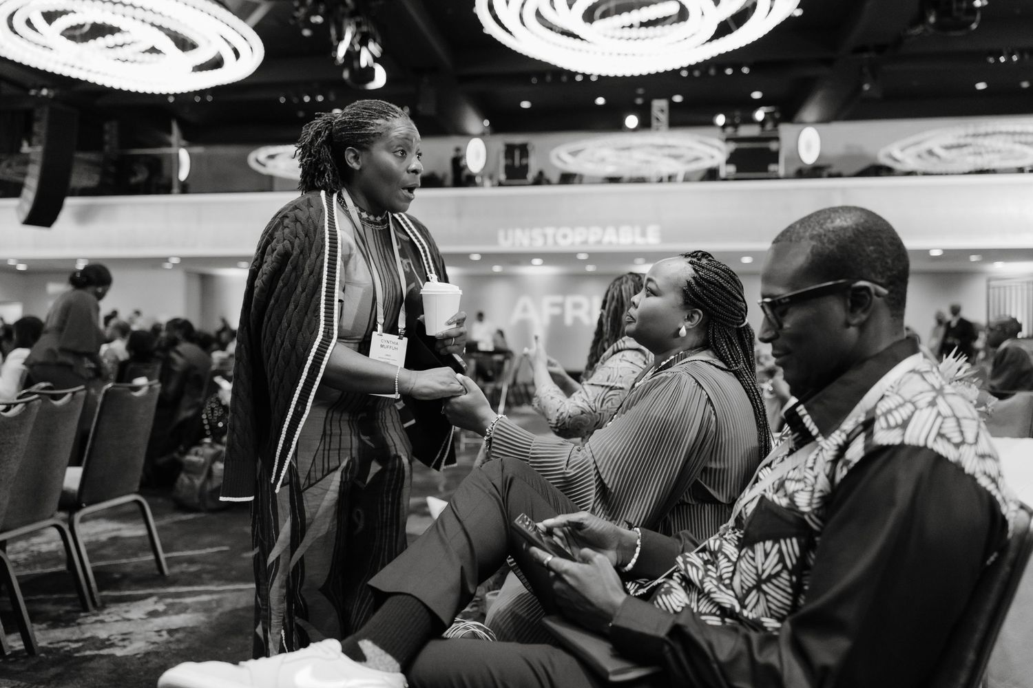 A black and white candid moment shows people engaged in conversation at an indoor event with circular lighting overhead.