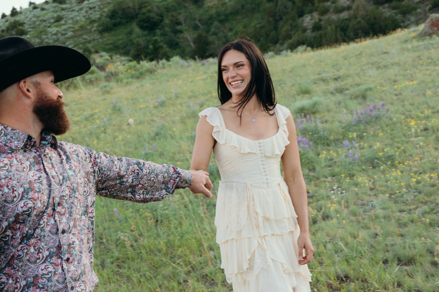 A couple stands together in a grassy meadow with wildflowers during a sunny afternoon.