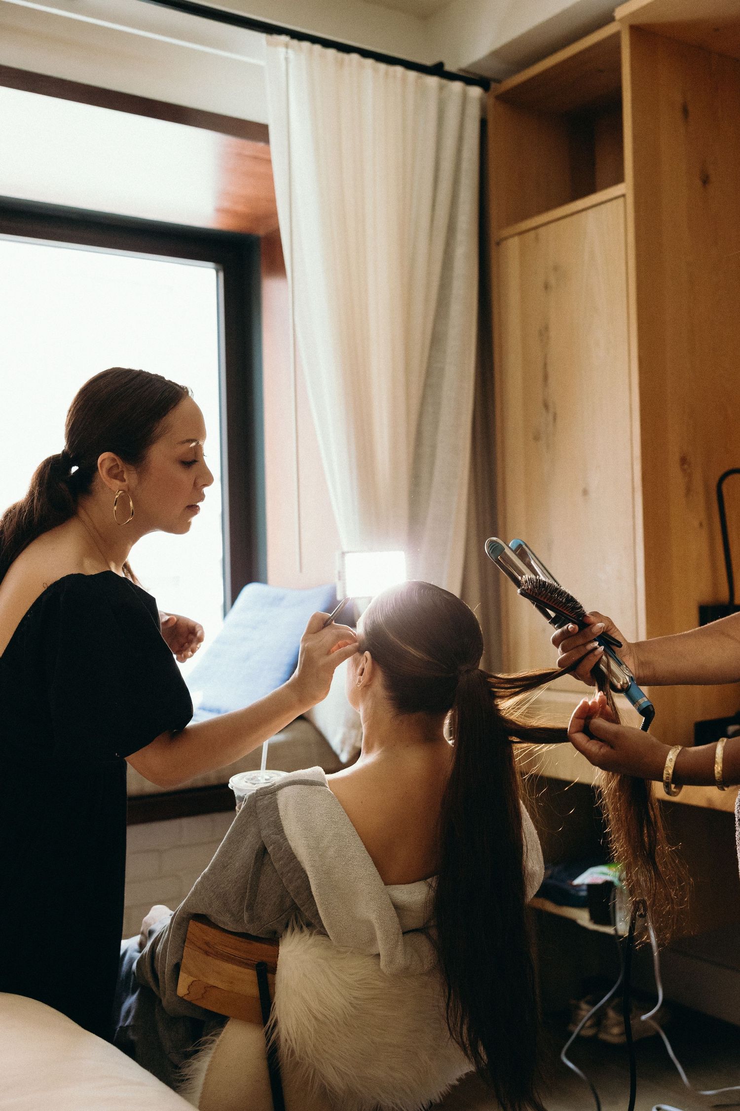 A hairstylist works on styling long dark hair in a sunlit room with wooden accents and natural lighting.