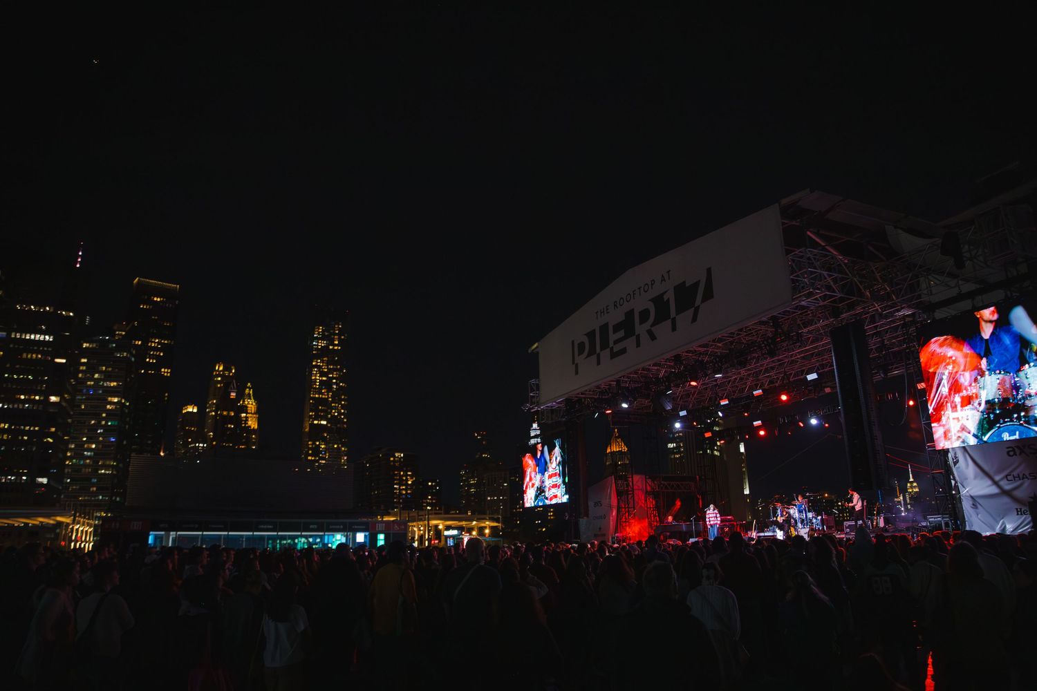 Nighttime cityscape view with concert stage and crowd silhouettes in foreground.