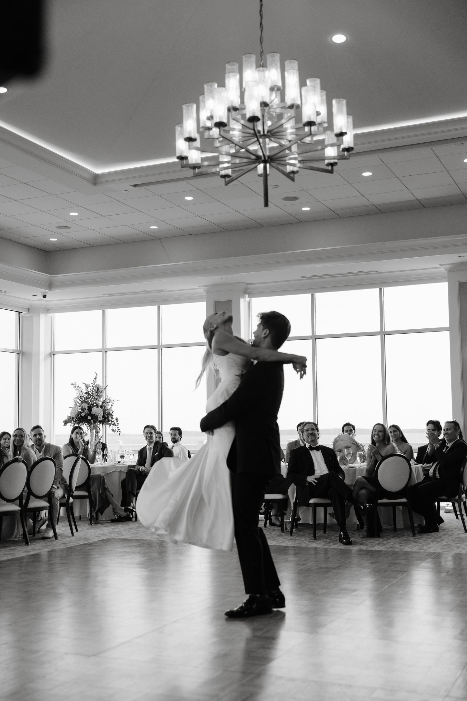 Black and white photo of couple's first dance in an elegant ballroom with large windows.