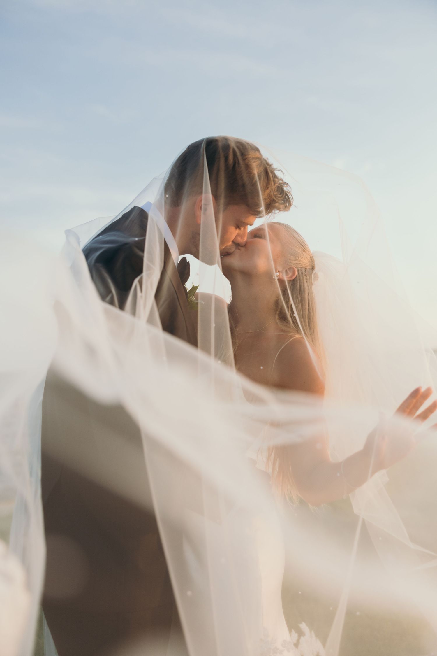Artistic wedding portrait through flowing white veil against sunset sky.