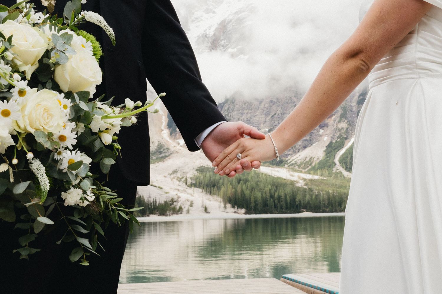 Close up of newlyweds holding hands with white floral bouquet against mountain lake backdrop.