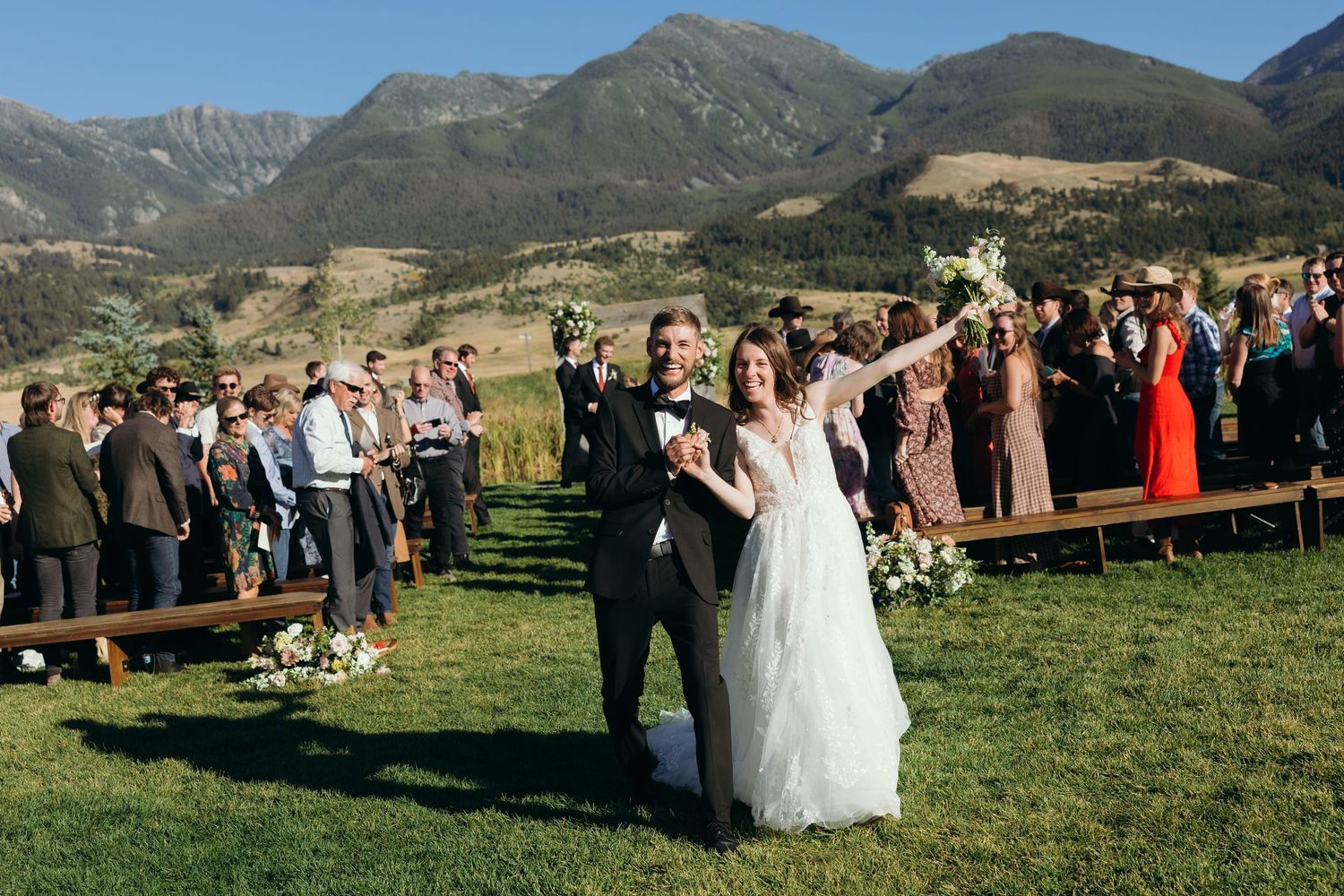 Outdoor wedding ceremony with mountain backdrop and guests seated on wooden benches.
