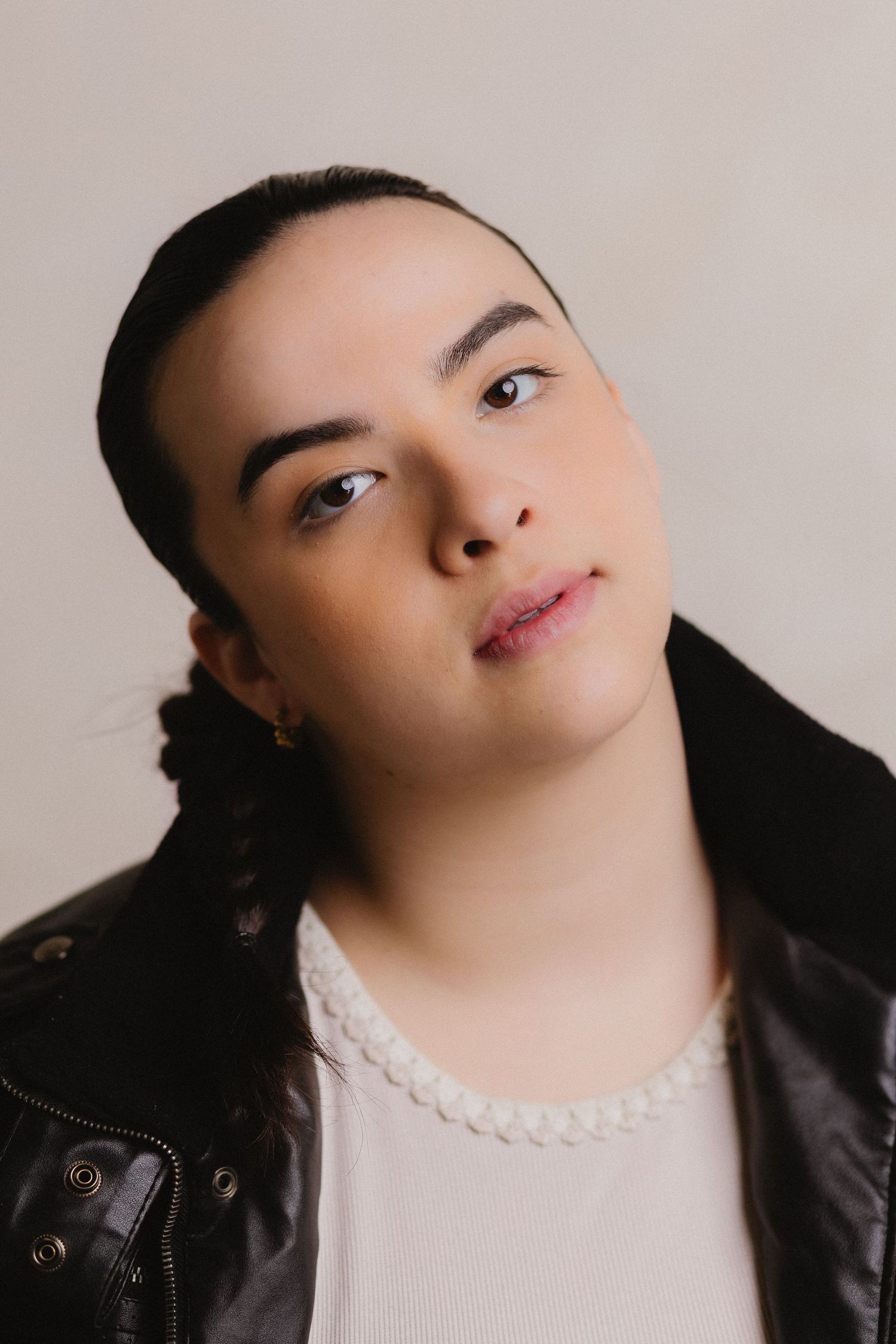 A person in a black leather jacket and white top poses with dramatic head tilts against a neutral background.