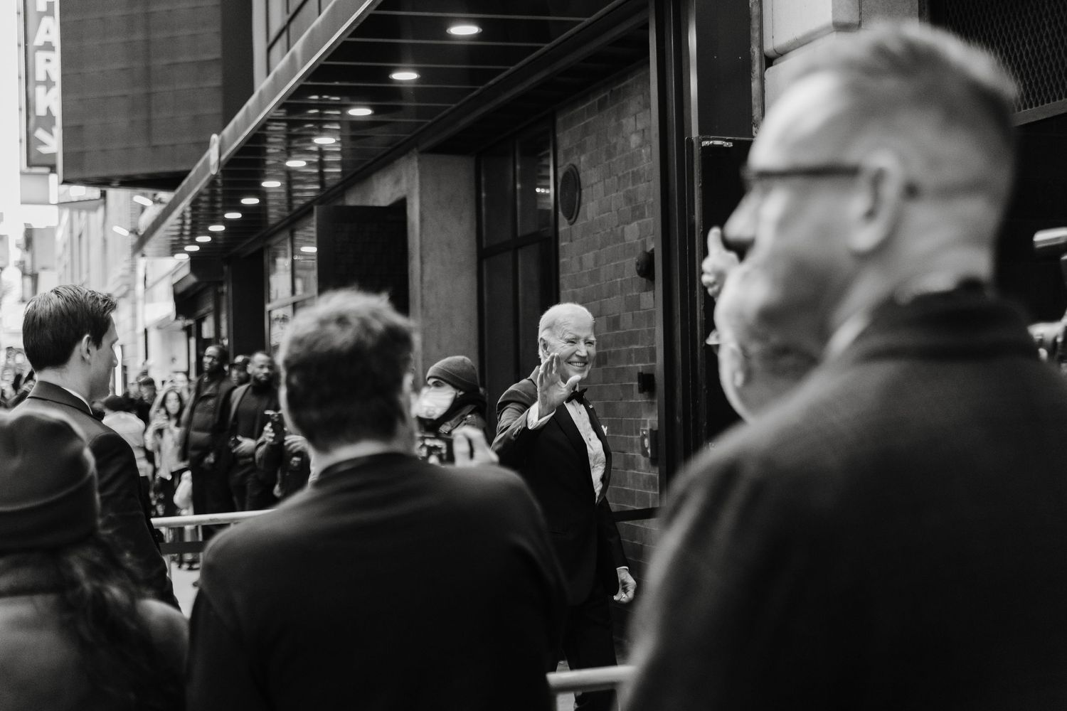 Black and white street photo of a crowd outside a building, with a man raising his hand in a saluting gesture.