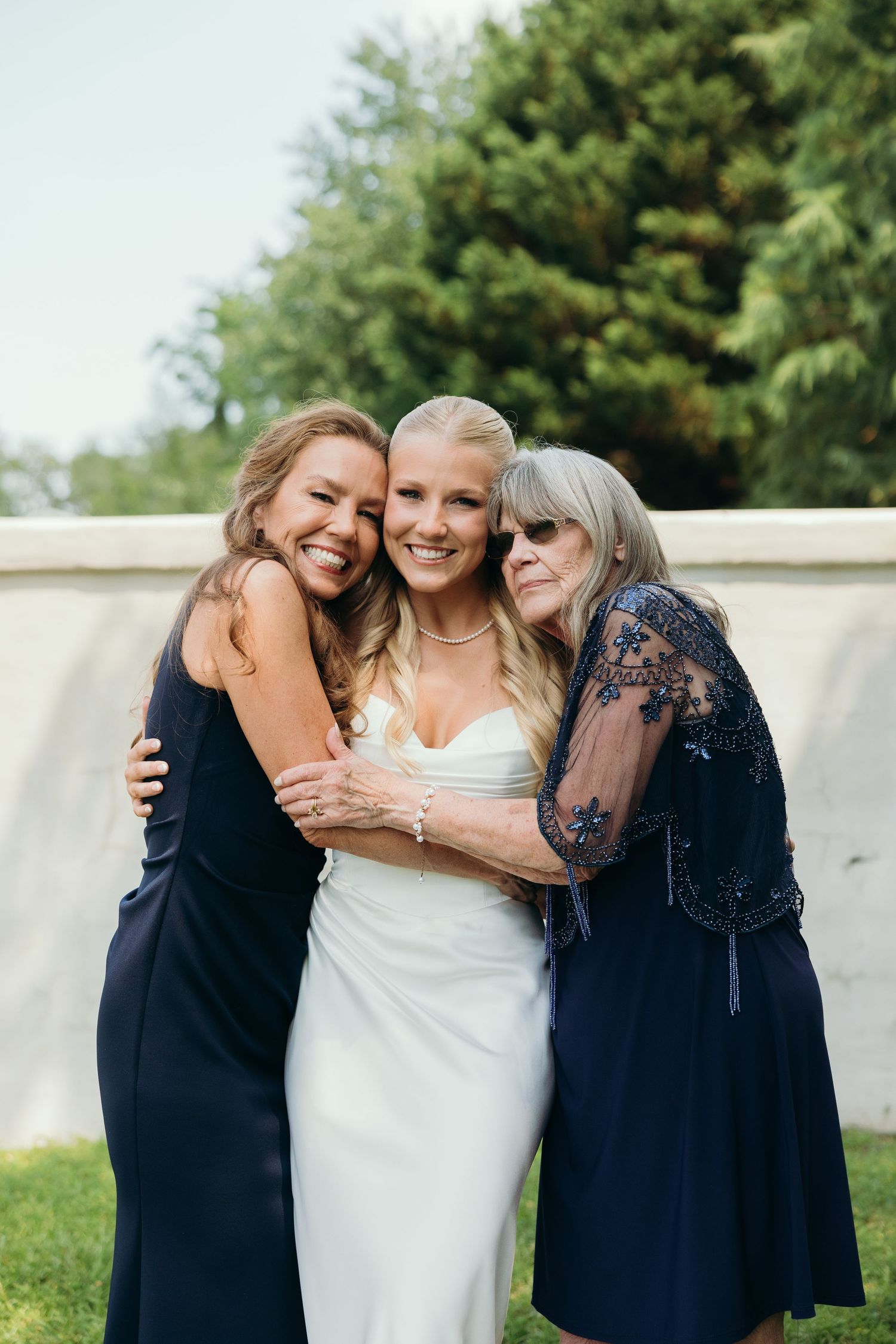 Three women share an emotional embrace outdoors during a wedding celebration.