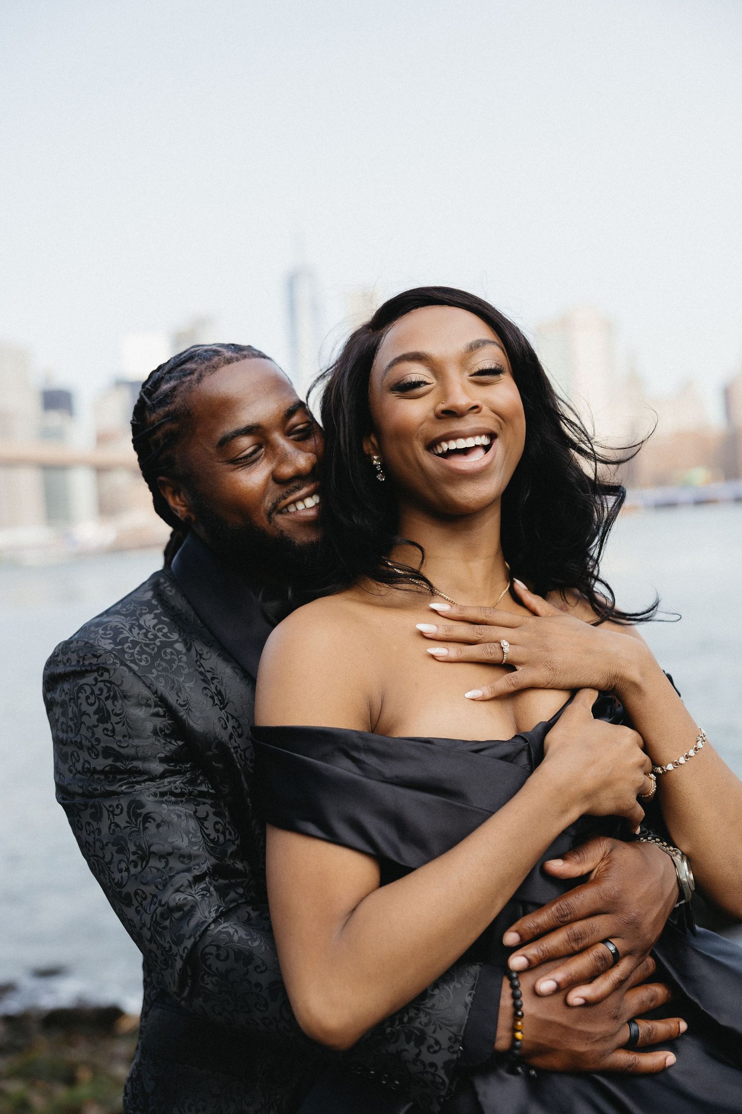 Couple laughing and embracing joyfully by the waterfront with city skyline in background.