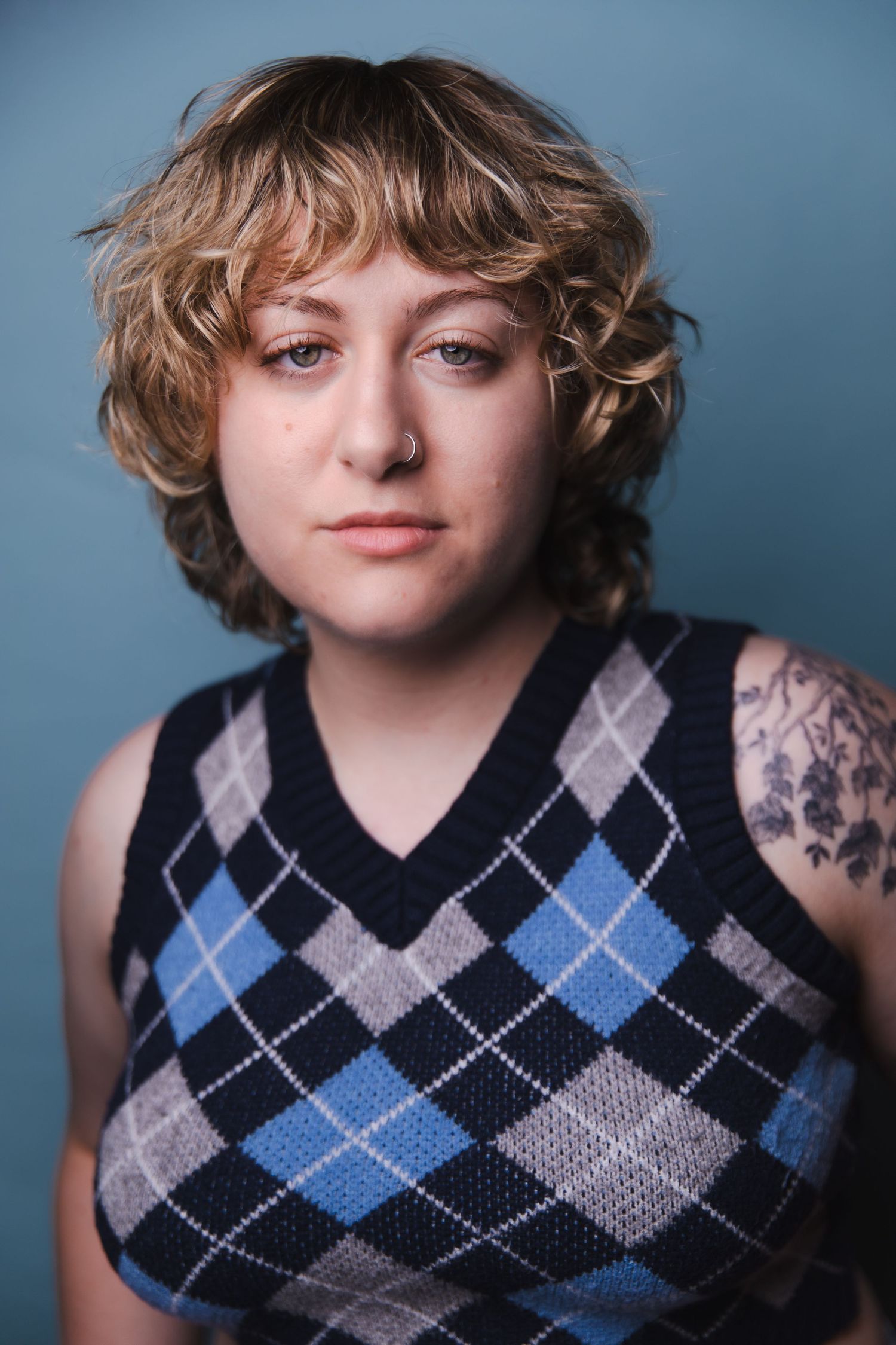 Person wearing a blue and gray argyle sweater vest with visible arm tattoos against a blue background.