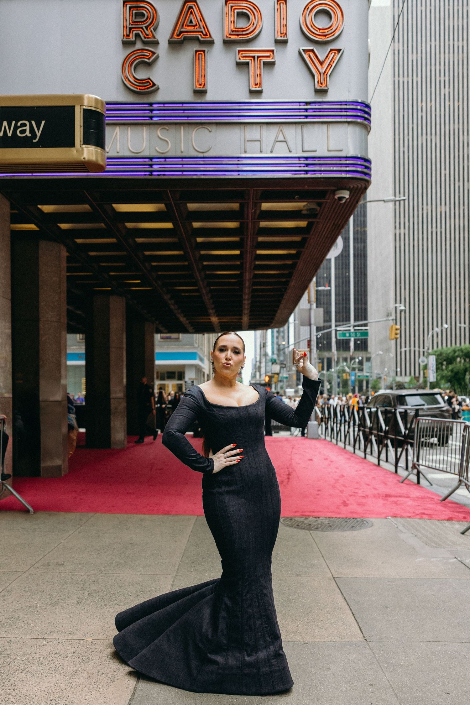 A glamorous photo of someone in a black mermaid gown posing outside Radio City Music Hall in New York City.