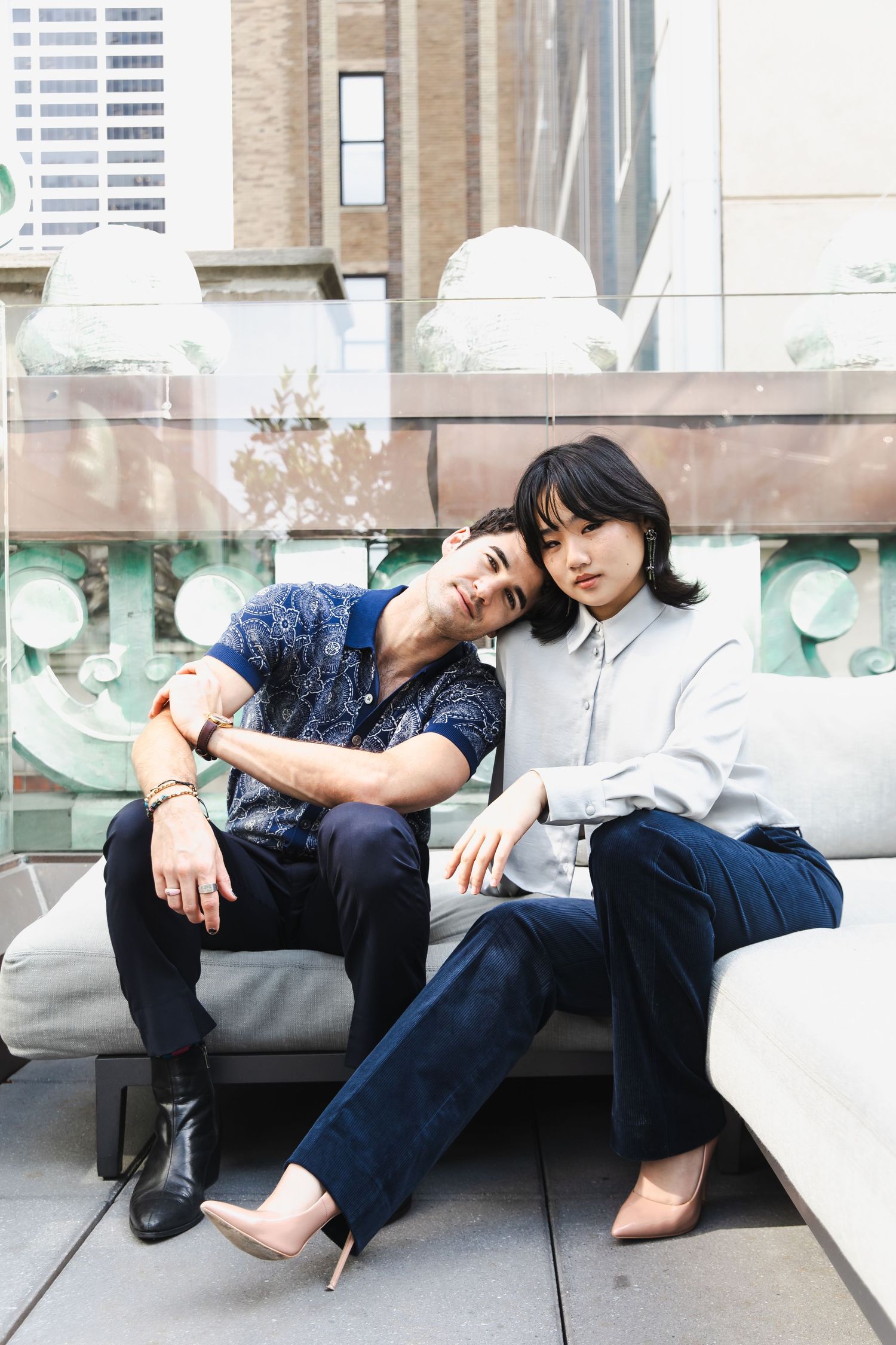 A man and woman sit closely together on an outdoor rooftop sofa, posing for a portrait in an urban setting.
