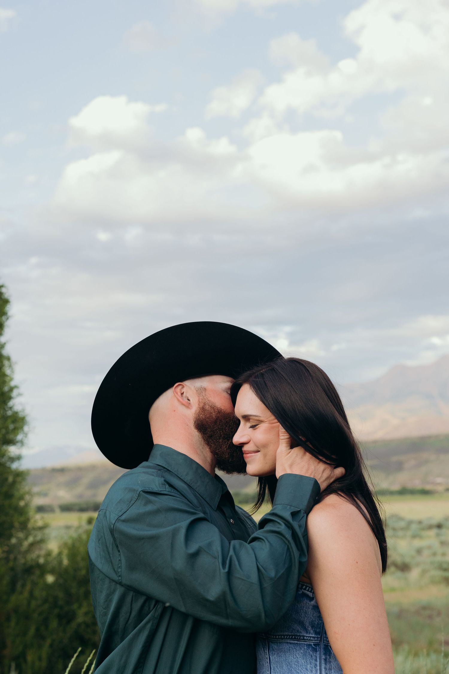 A romantic couple embraces in front of a scenic mountain landscape on a cloudy day.
