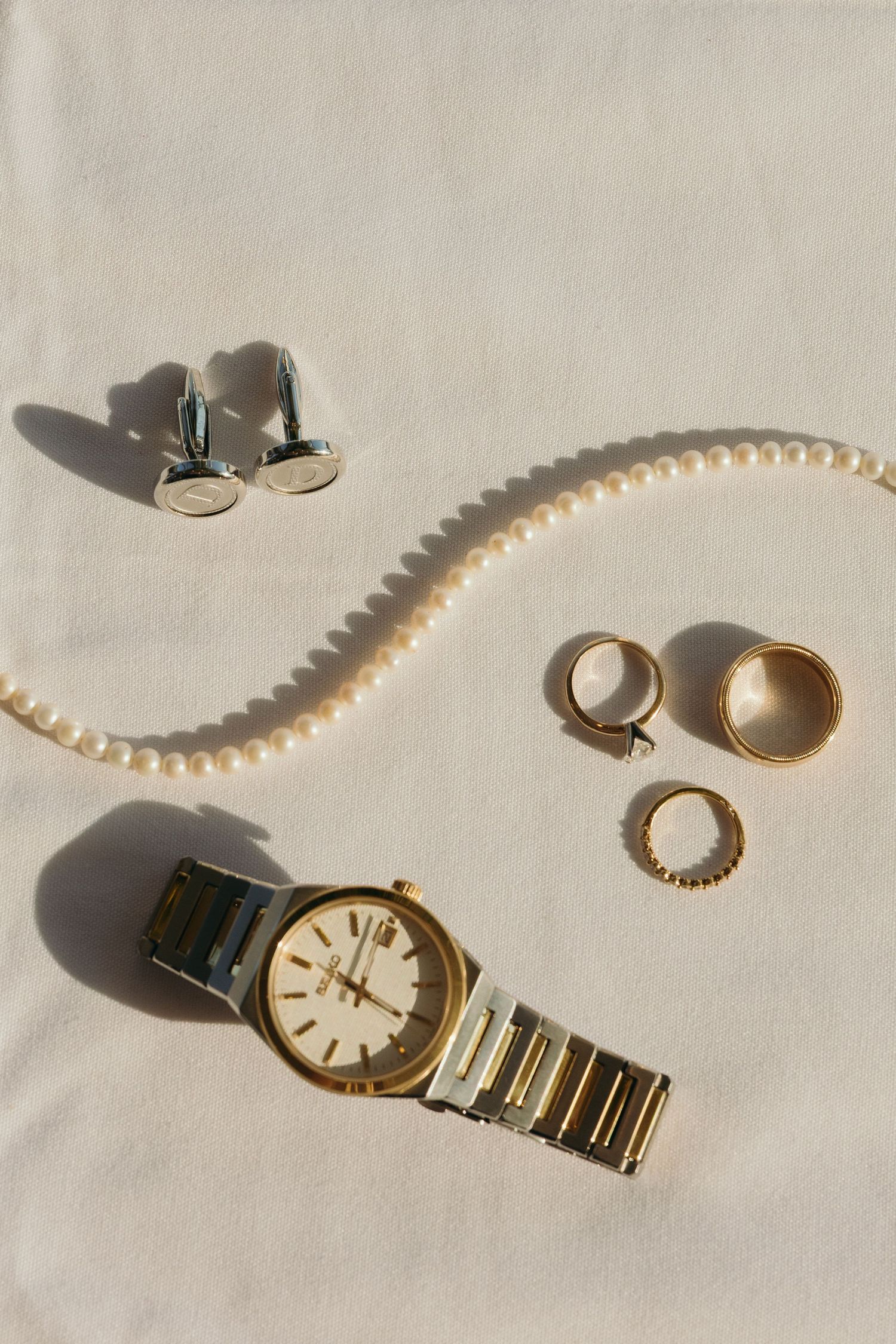 Wedding accessories including pearls, jewelry, and a gold watch arranged on white fabric.