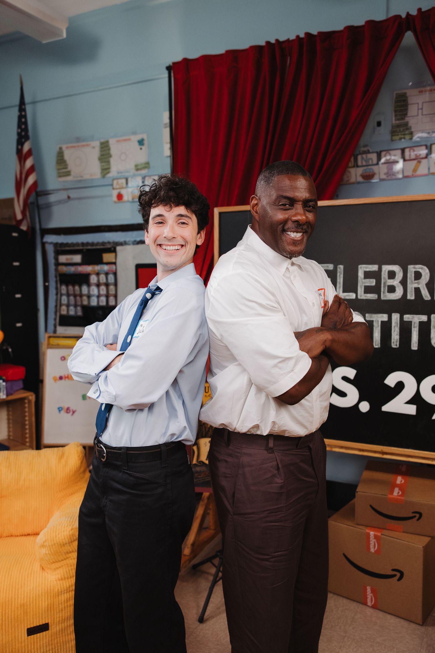 Two smiling men in white shirts stand back-to-back with arms crossed in a decorated classroom near a chalkboard.