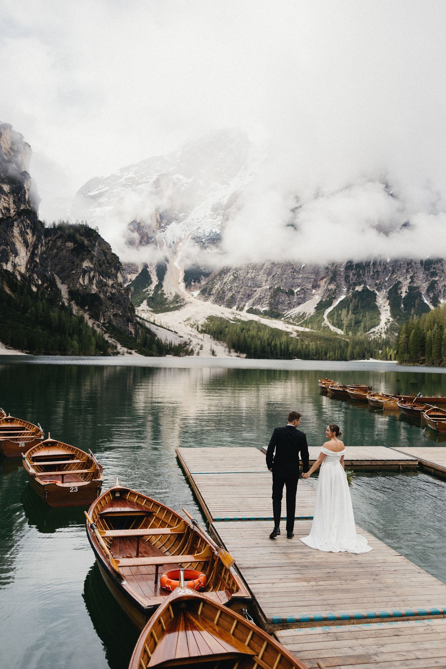 Wedding couple standing on a wooden dock with boats at a misty mountain lake.