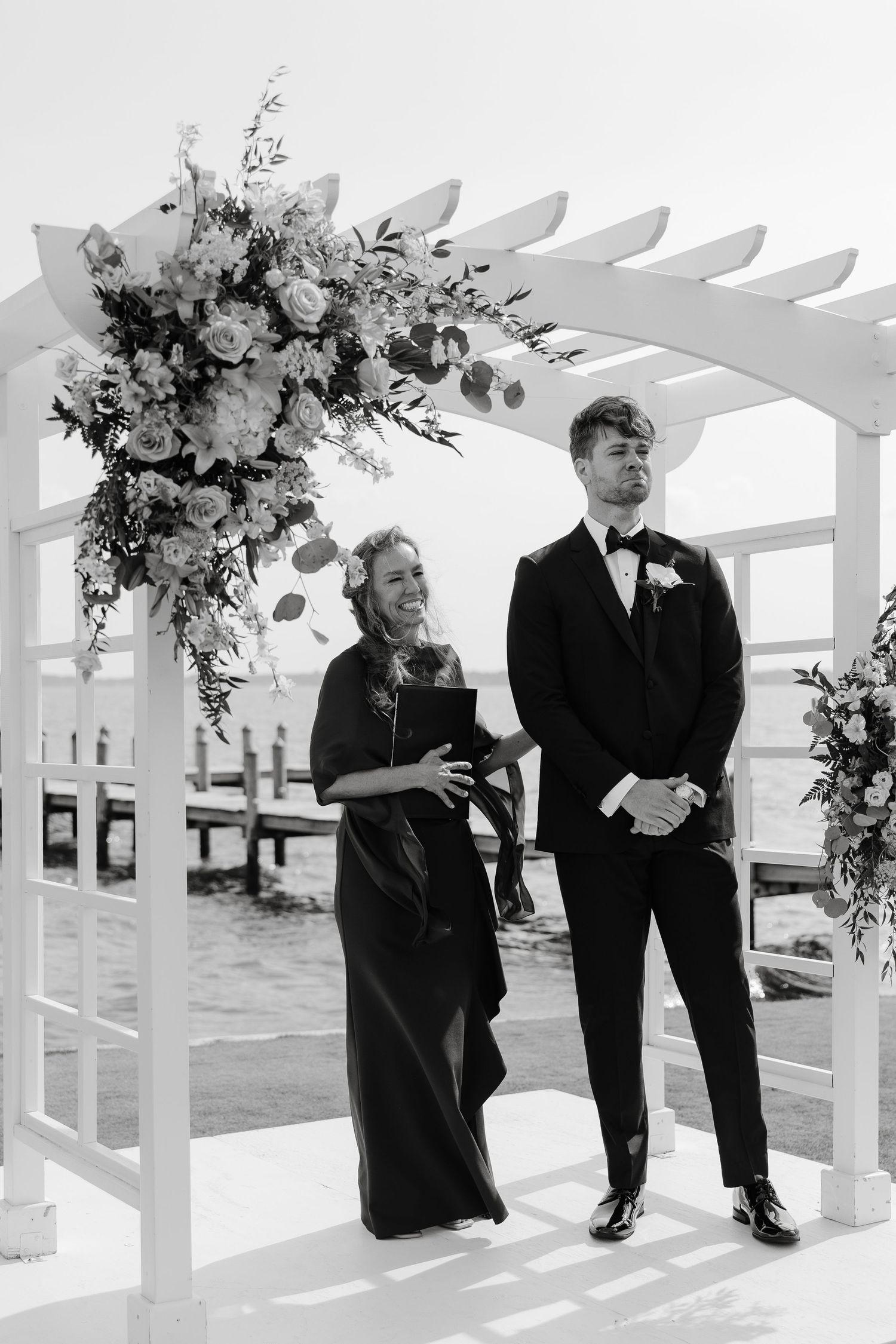 Wedding party members in formal attire laughing under white pergola decorated with flowers.