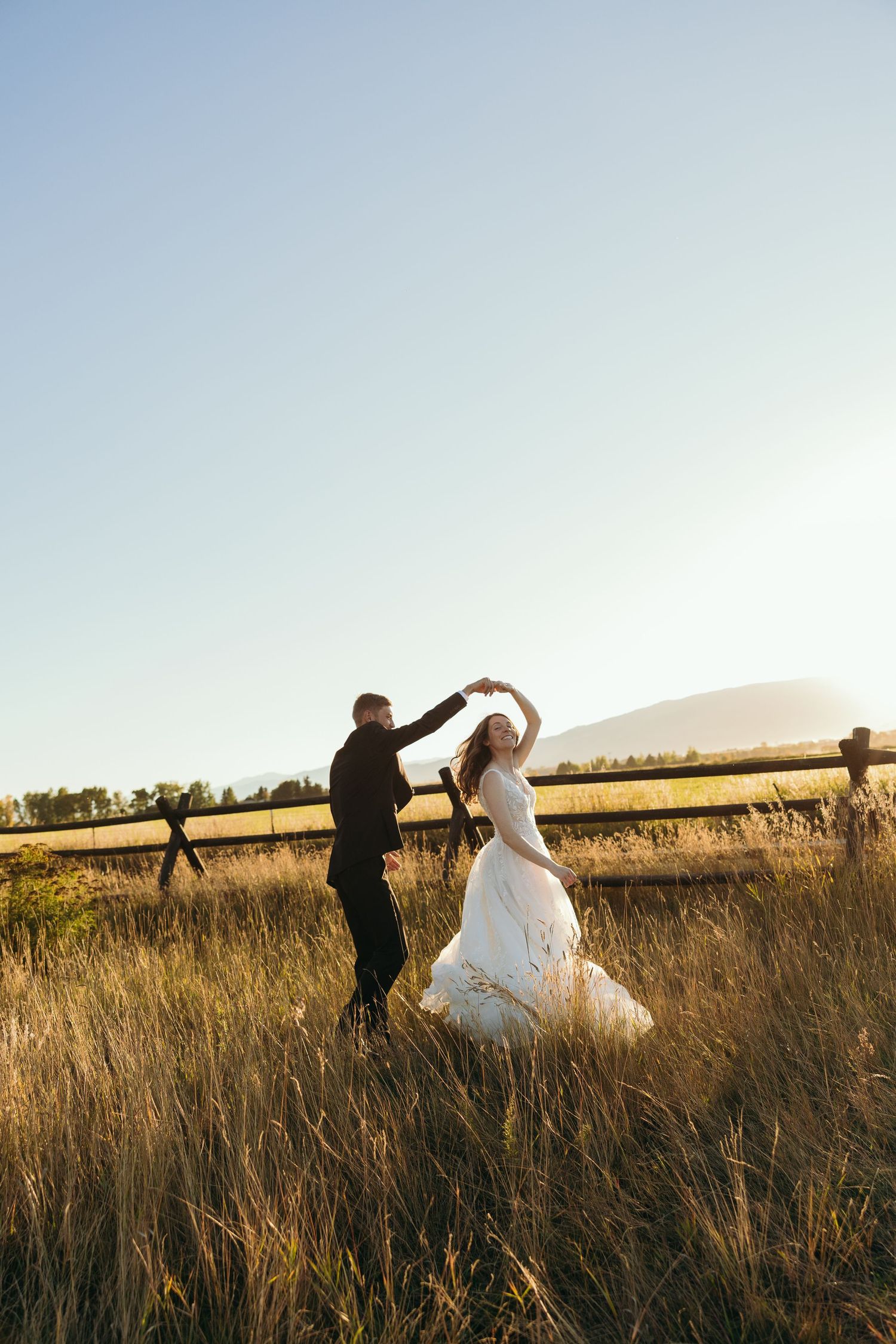 Couple dances in a golden field at sunset with mountains in the background.