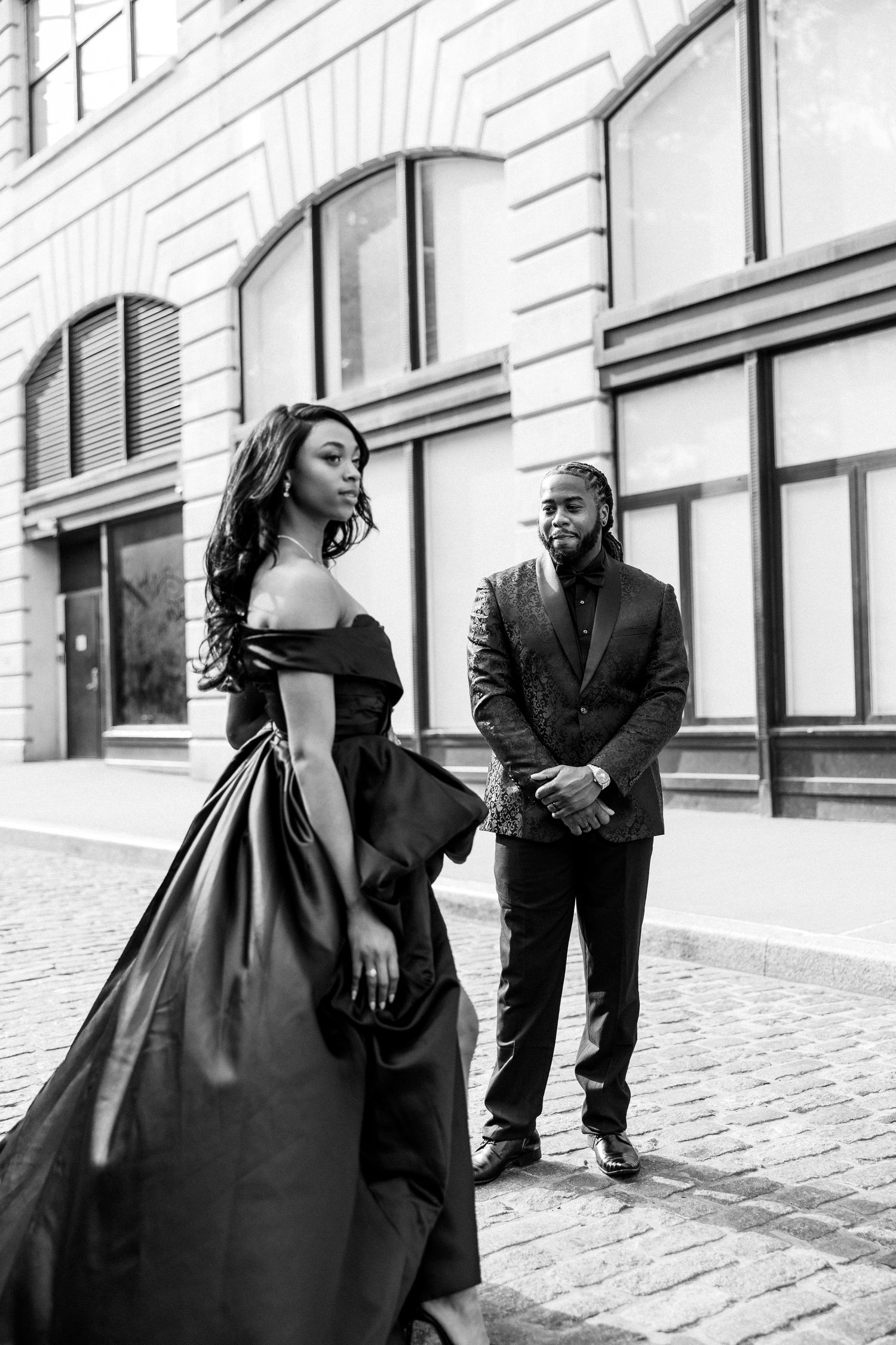 Playful engagement photos of couple in formal wear on urban cobblestone street.