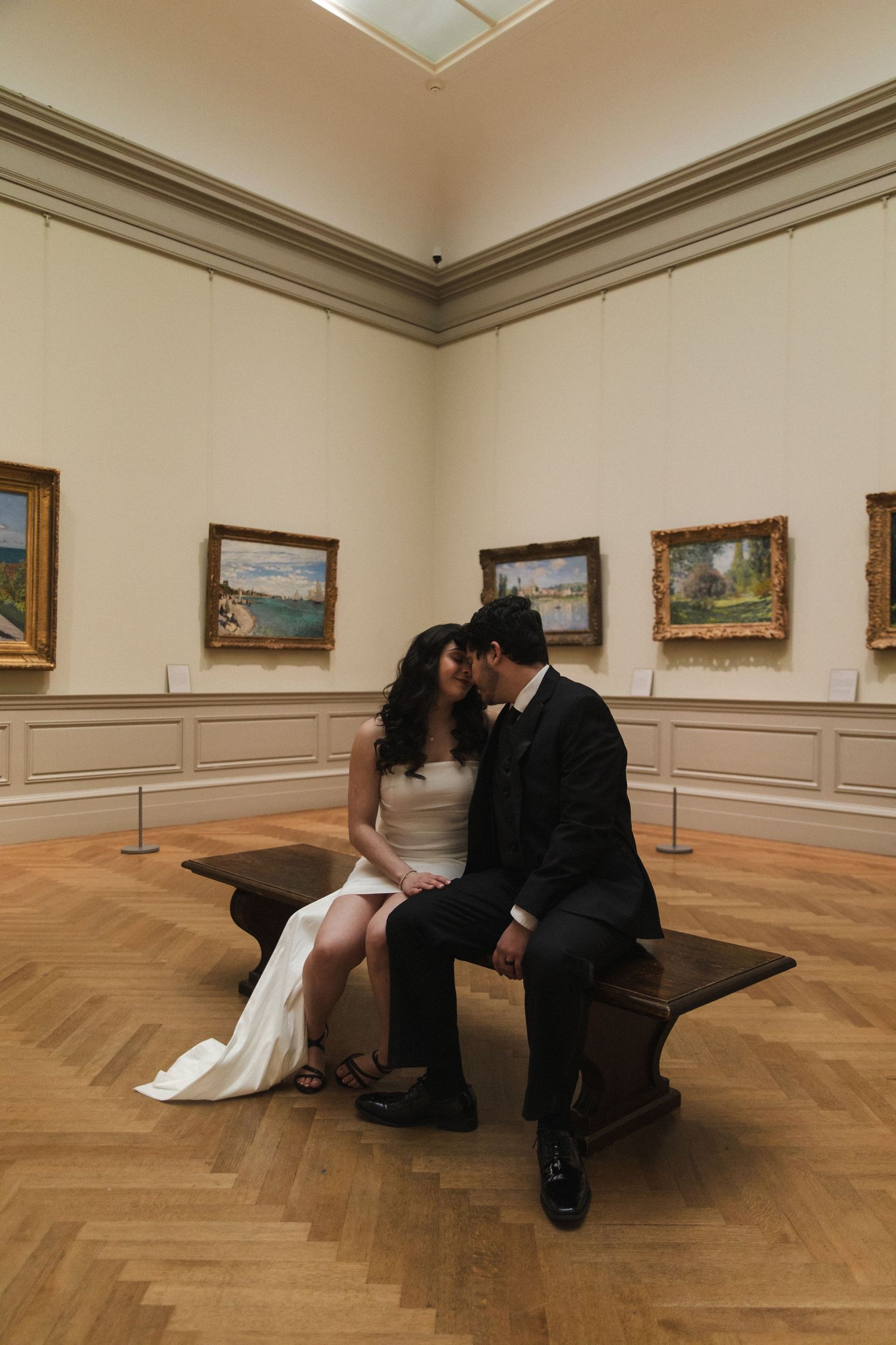 Wedding couple sitting close together in an art gallery surrounded by framed paintings.