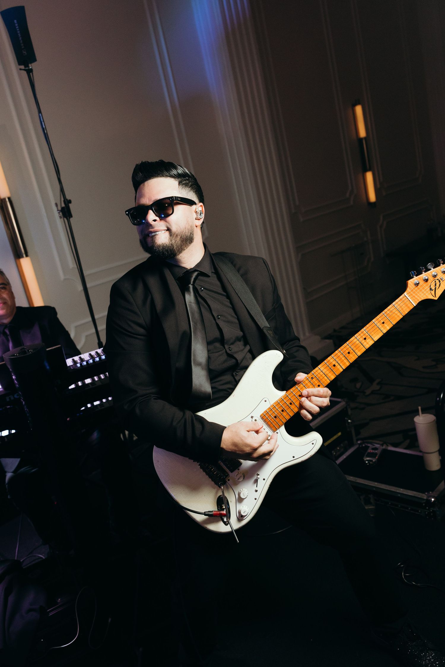 Guitarist in black attire playing a white electric guitar during a dimly lit performance.