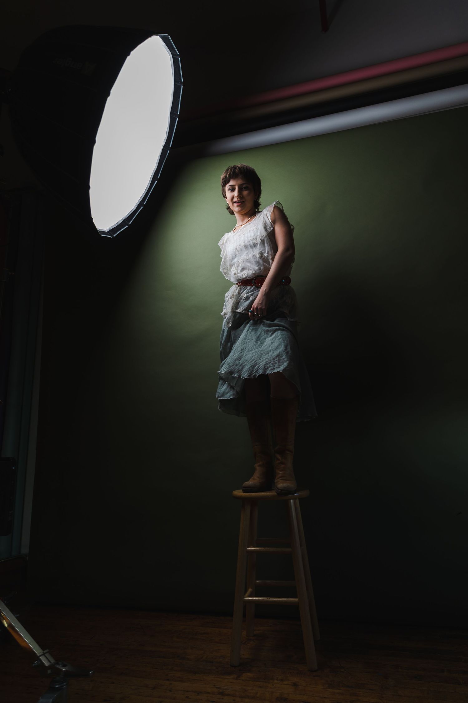 Studio lighting setup with person posing on wooden stool against dark background in dramatic lighting setup.