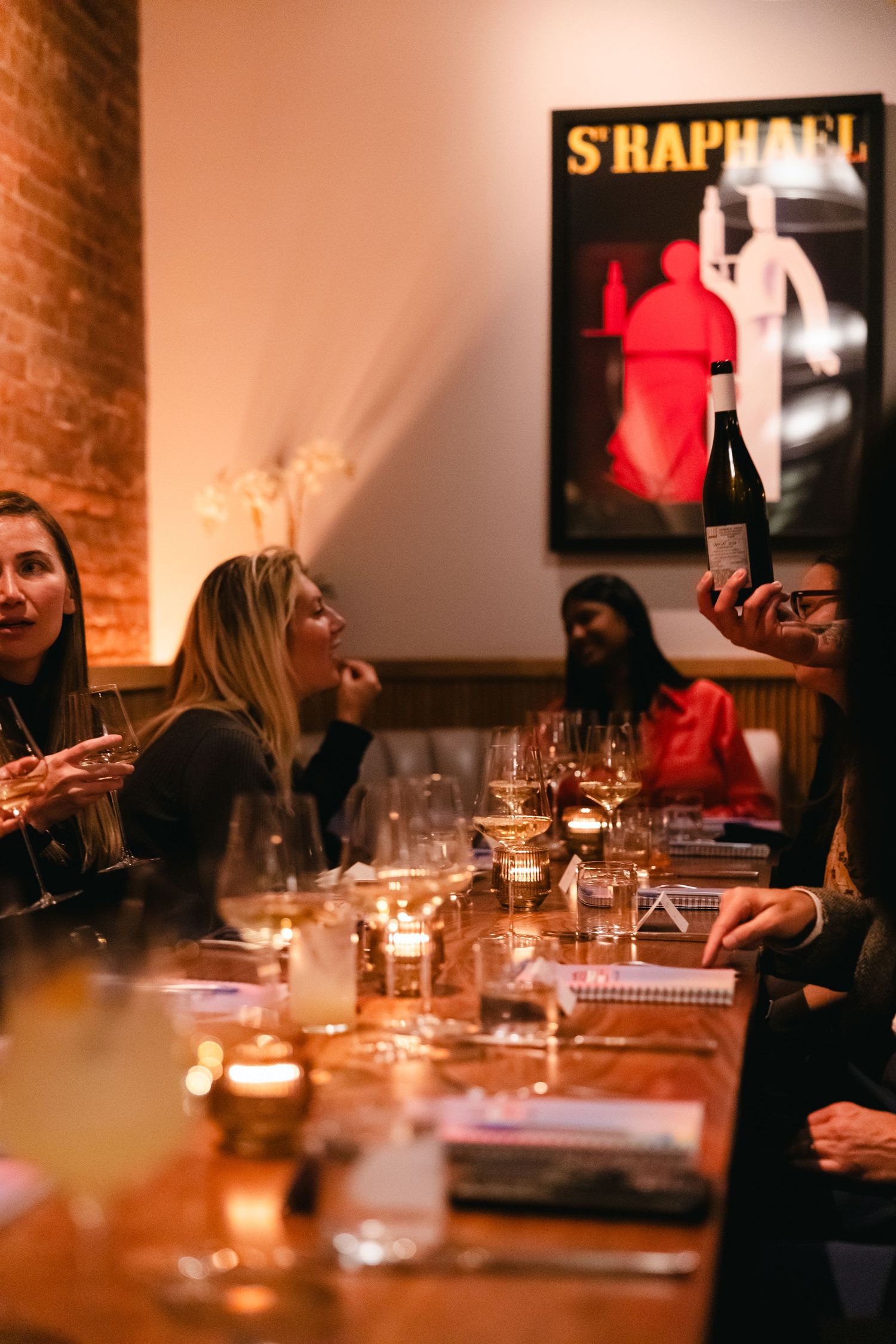 Dimly lit restaurant dining room with exposed brick walls and guests seated at a long communal table for dinner service.