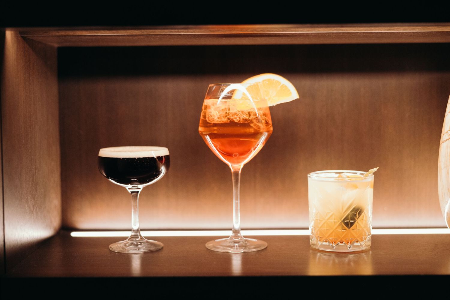 Various cocktail glasses filled with colorful drinks sit illuminated on a dark bar counter with warm ambient lighting.