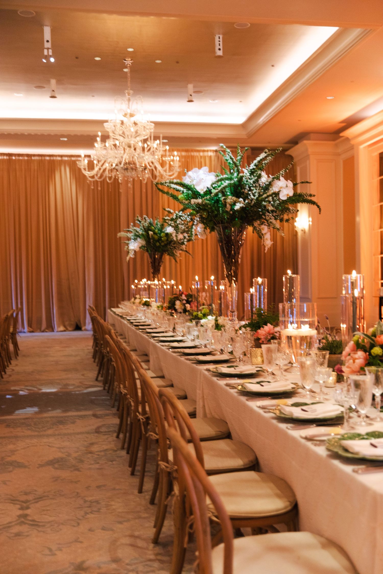 Long elegant dining tables set for a formal event with tall floral centerpieces and crystal chandeliers.