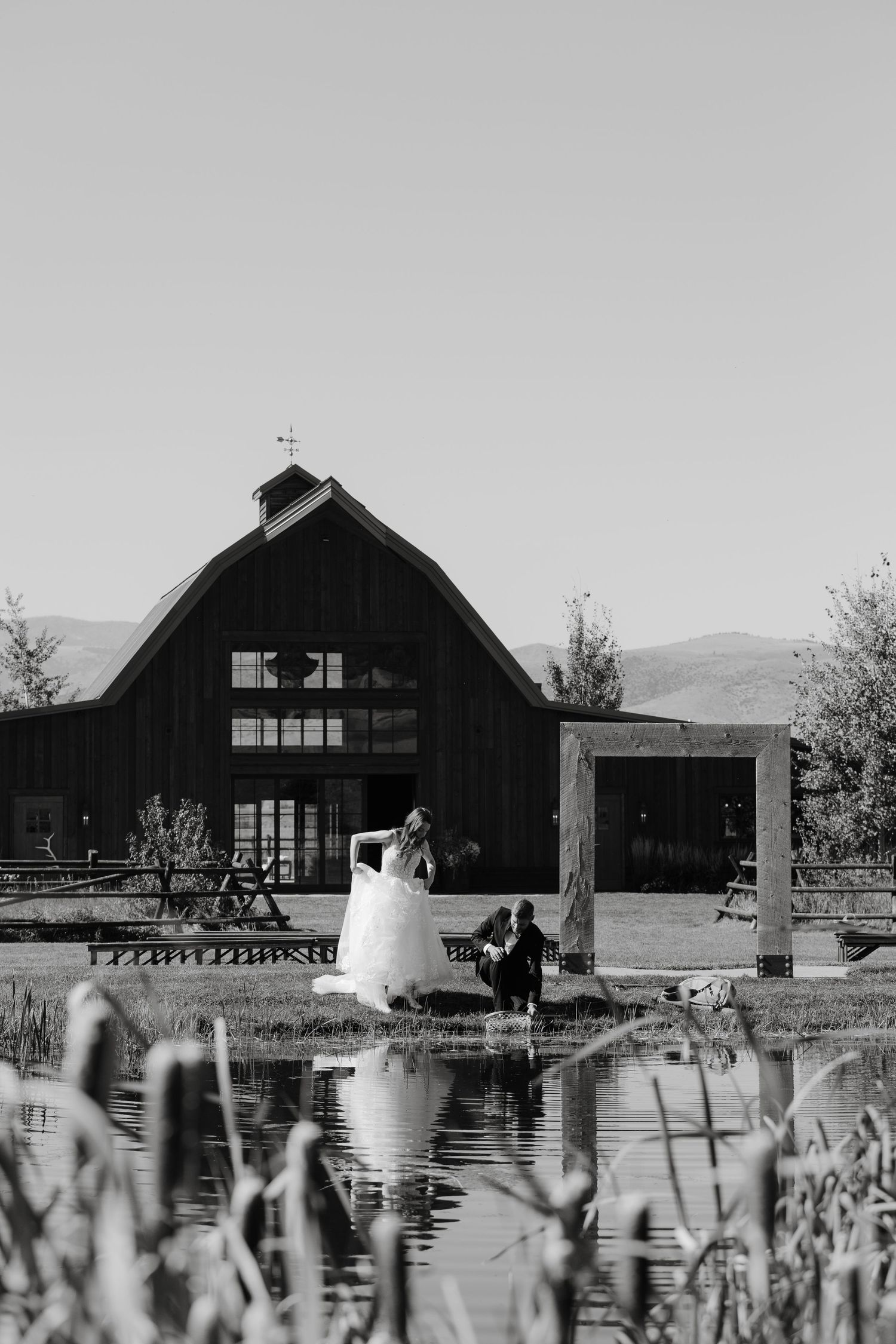 Black barn buildings reflect in still water with mountains in background.