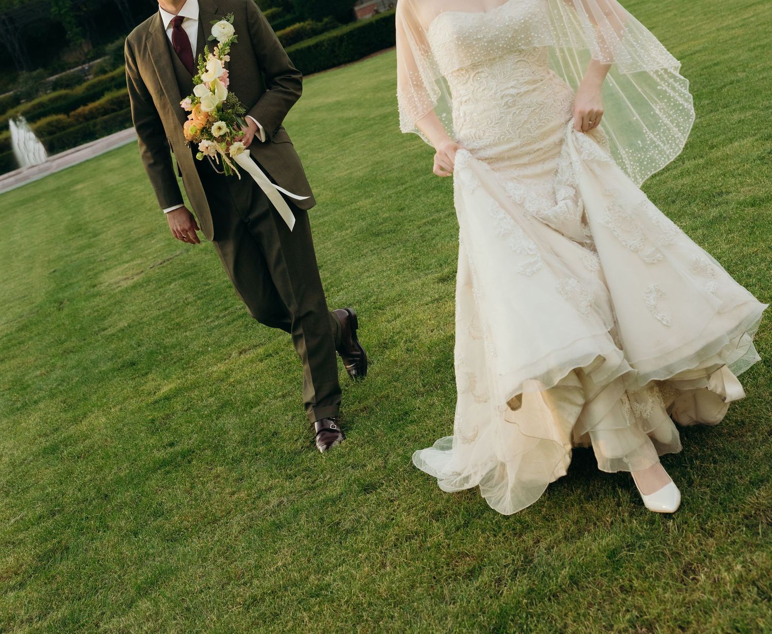 A cropped wedding photo showing the bottom half of a couple walking on grass, with flowing white dress and dark suit.