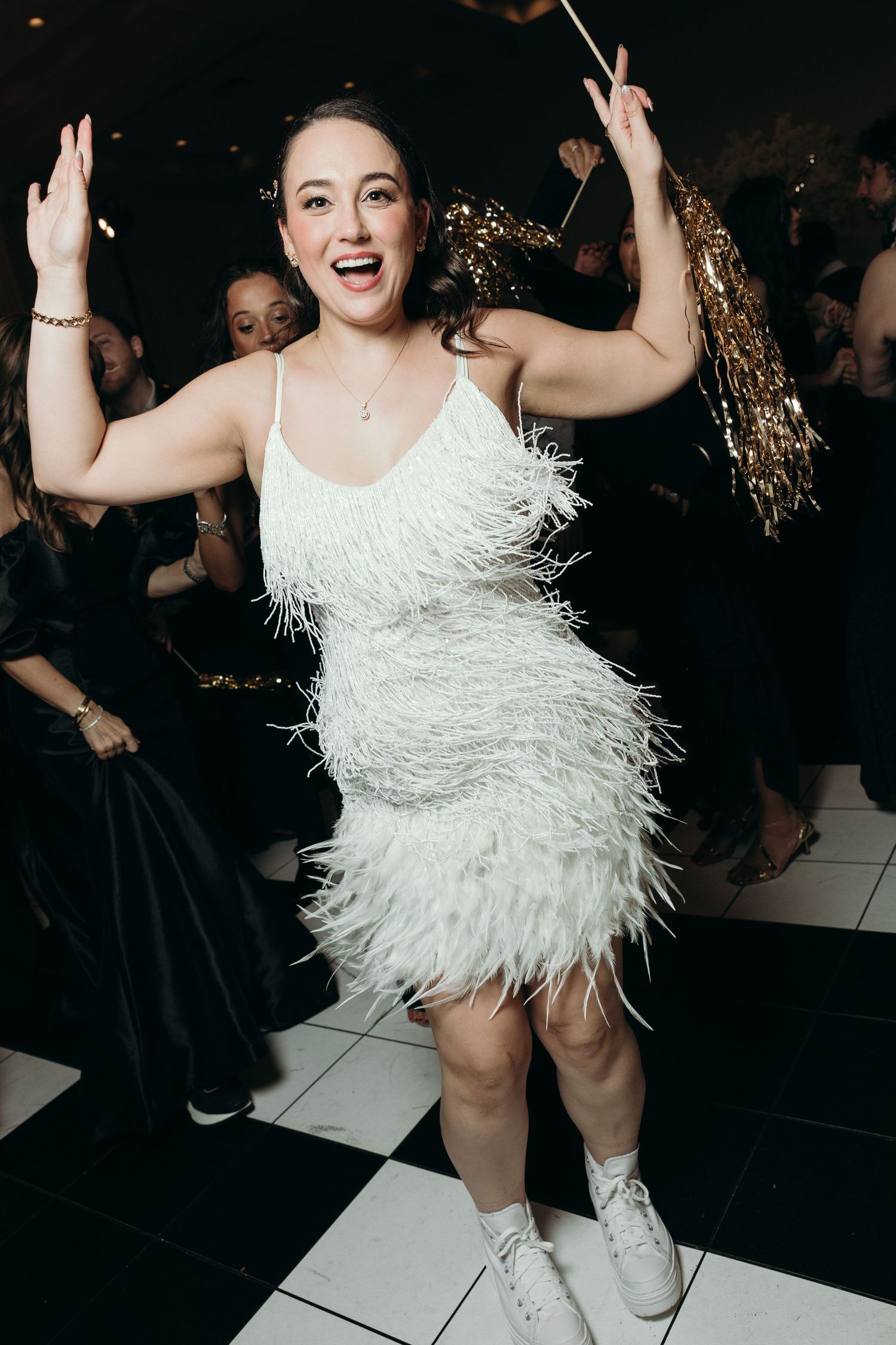Bride in white feathered party dress dances joyfully on checkered dance floor at reception.