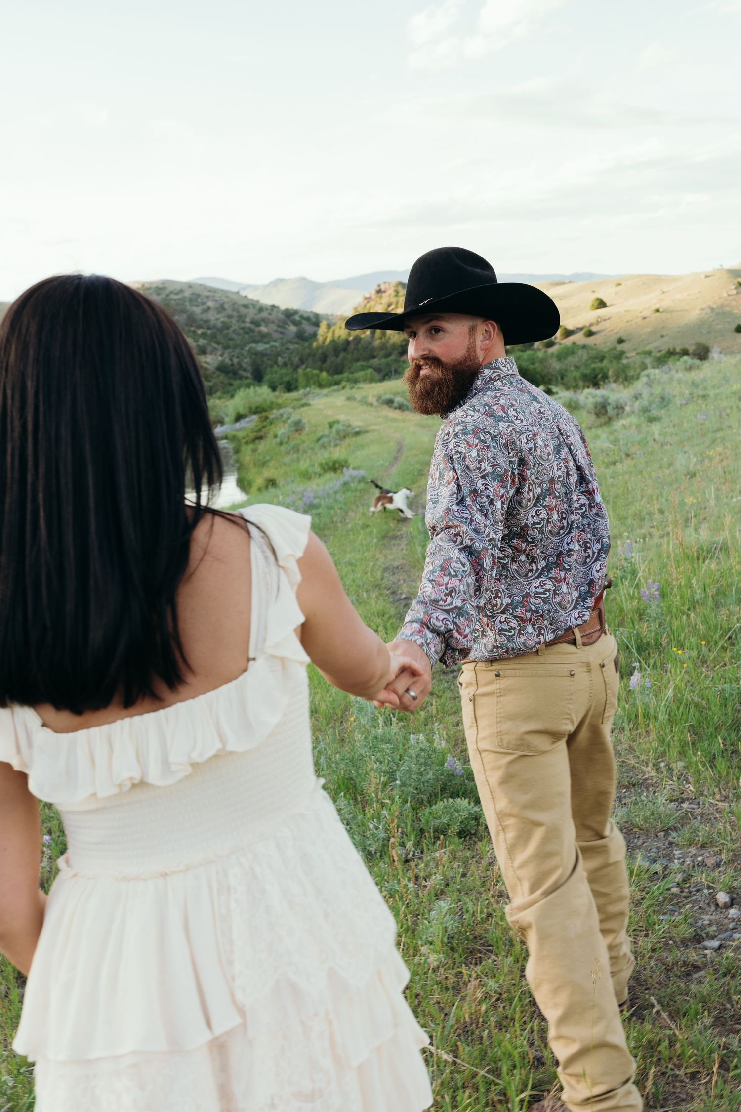 A couple walks hand in hand through a mountain meadow during sunset.
