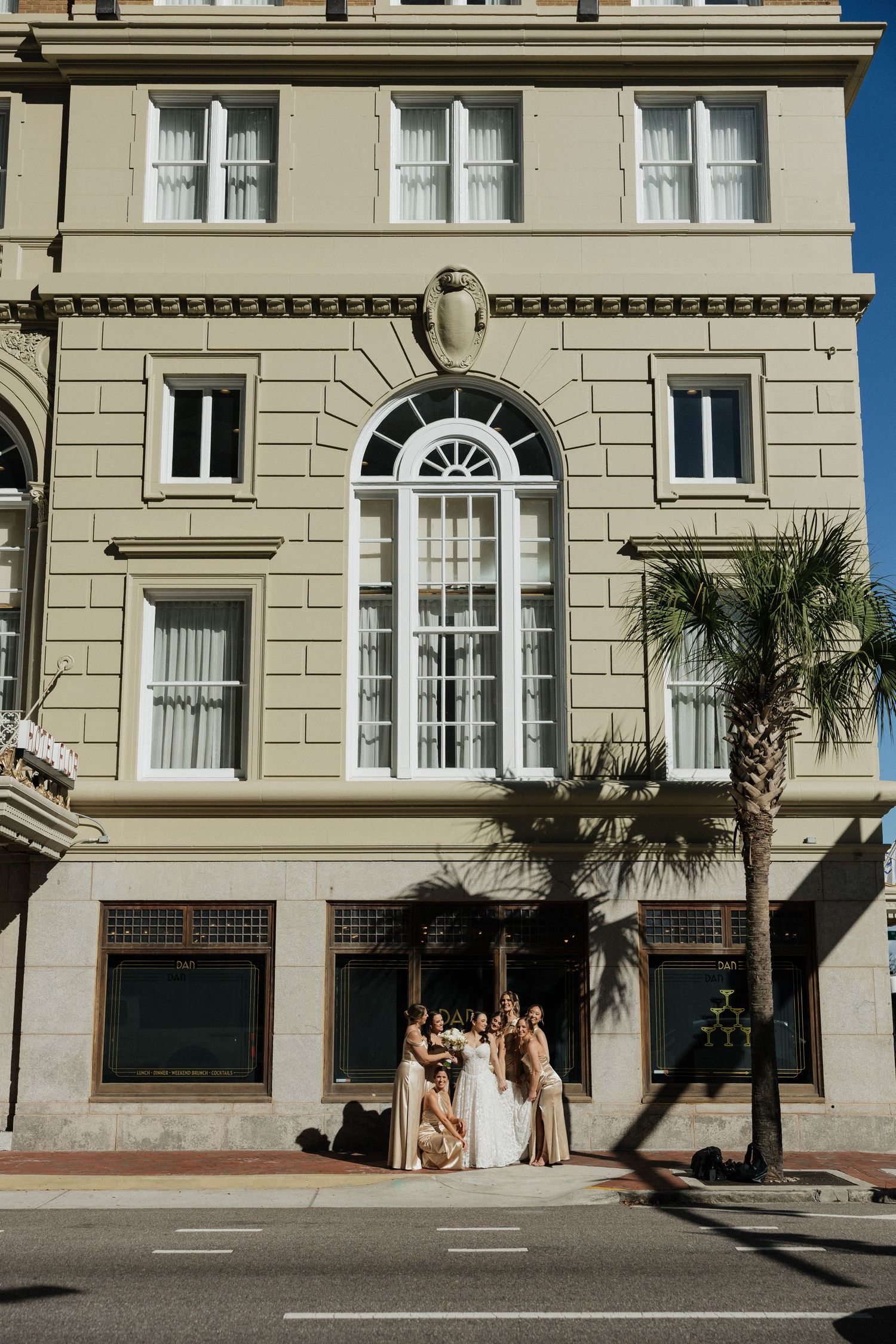 Wedding party gathered outside a historic beige building with palm trees and arched windows.