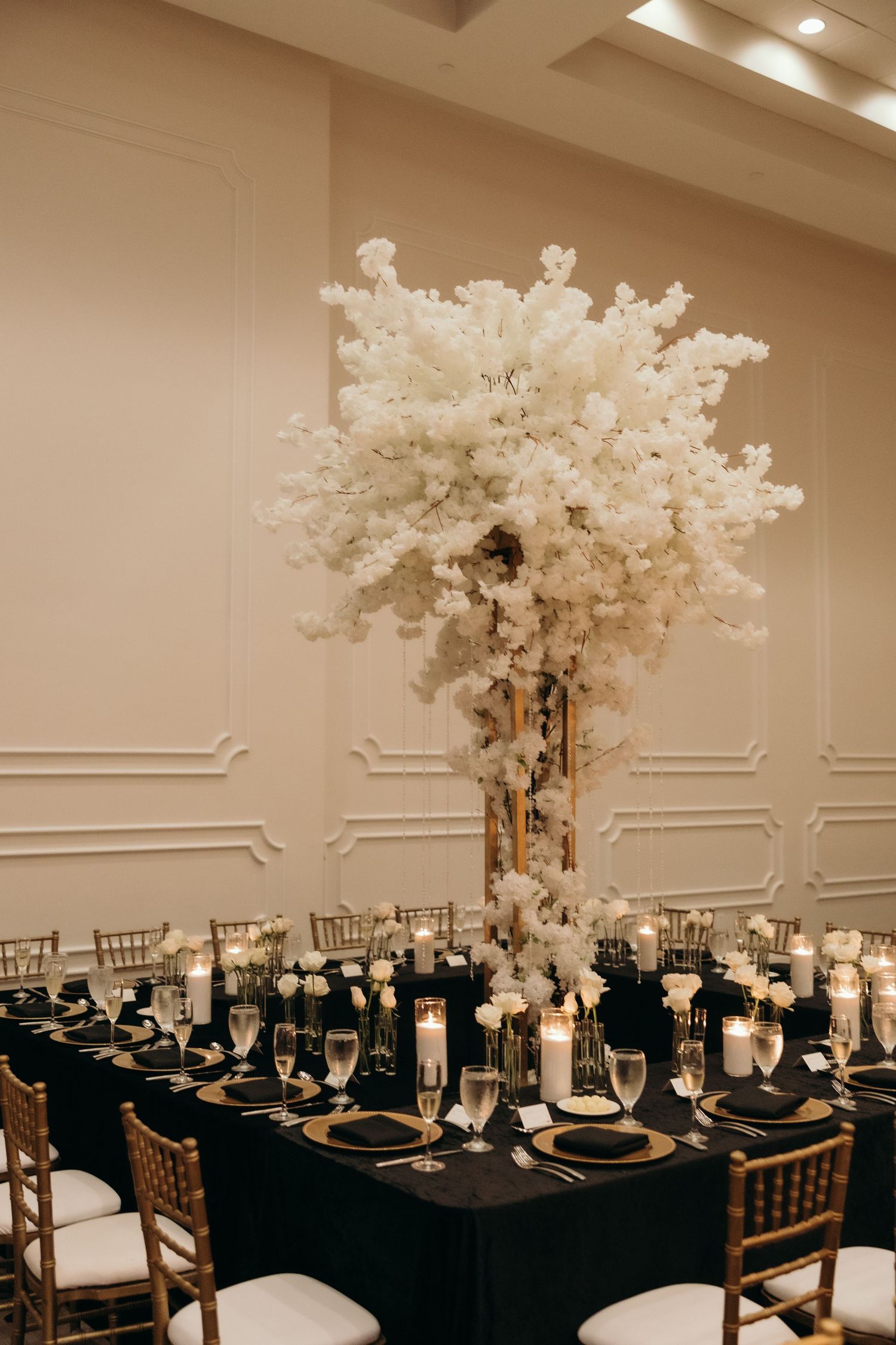 Elegant wedding reception tables set with black linens and tall white floral centerpieces in a luxurious ballroom setting.