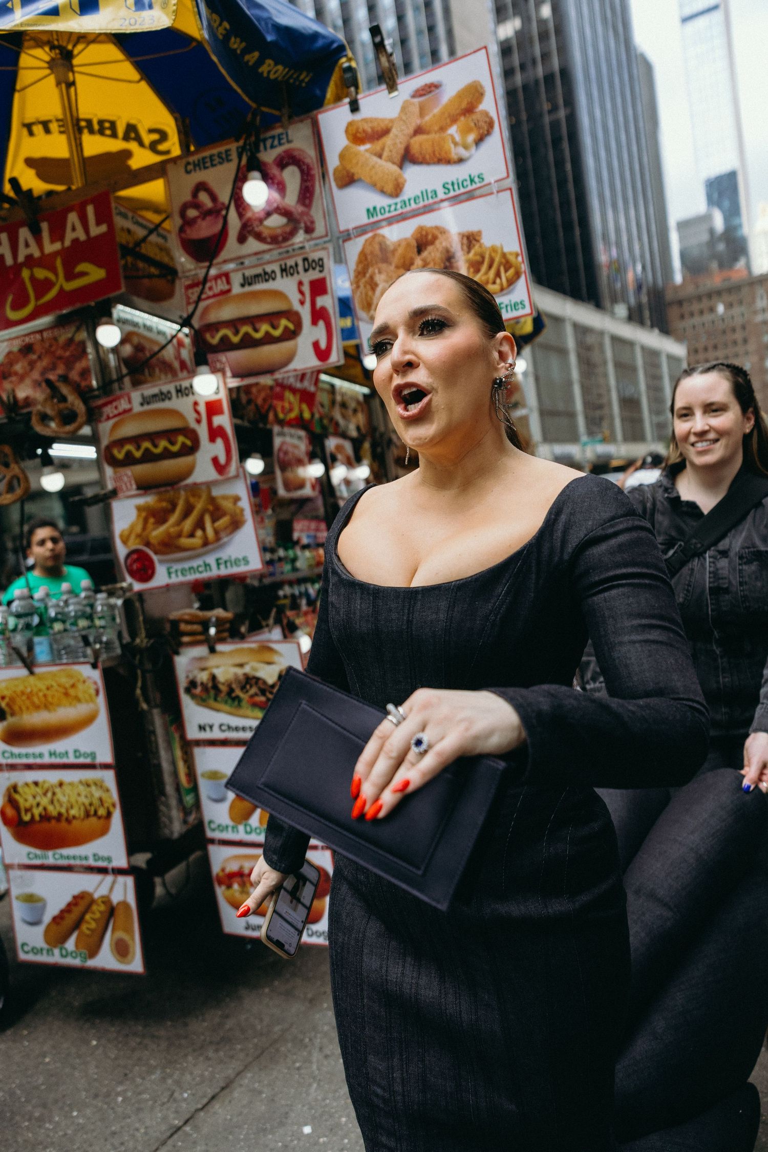 Jessica Vosk in a black sweater gestures expressively near food vendor carts on a city street.