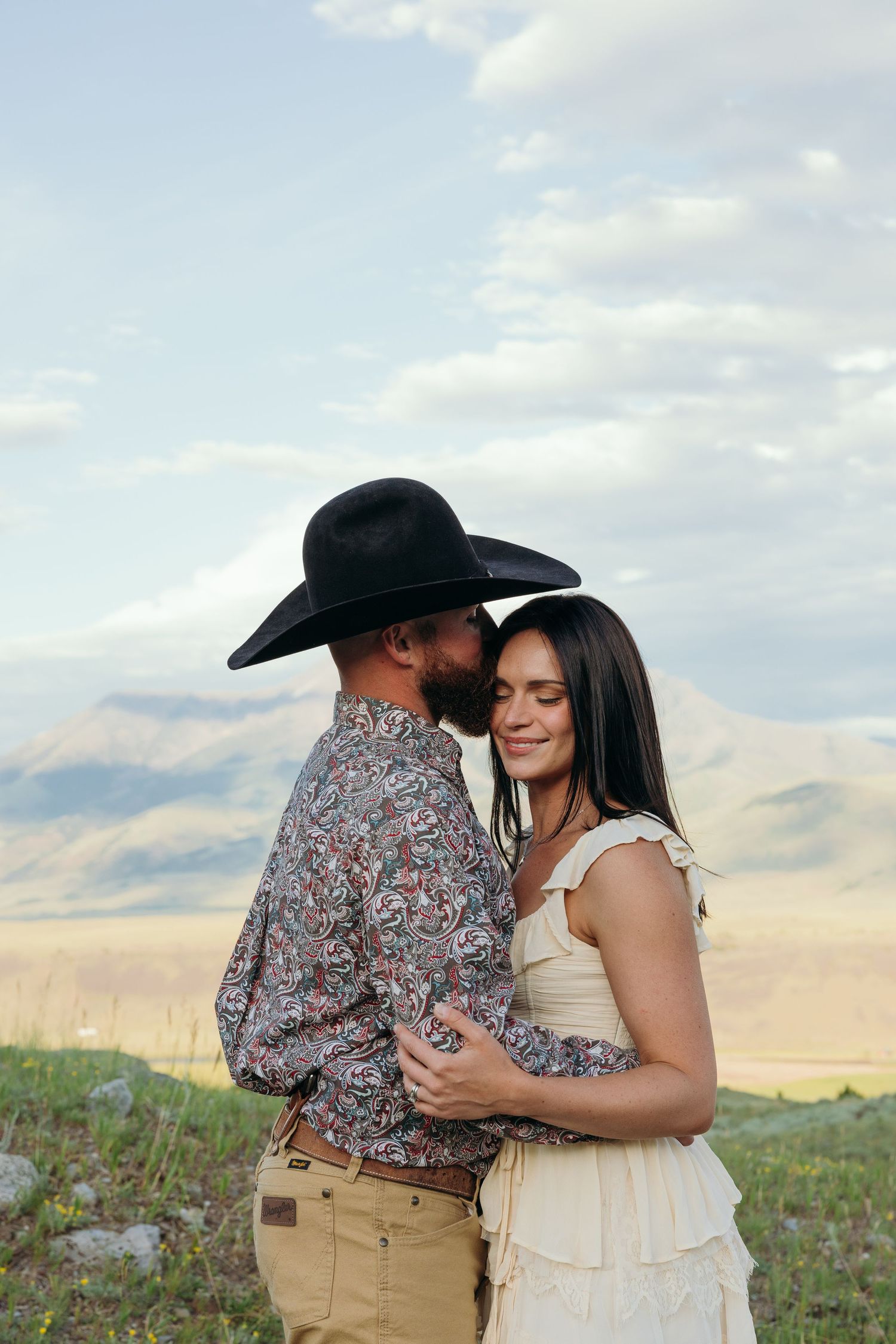 A couple embraces on a grassy hillside with mountains in the background.
