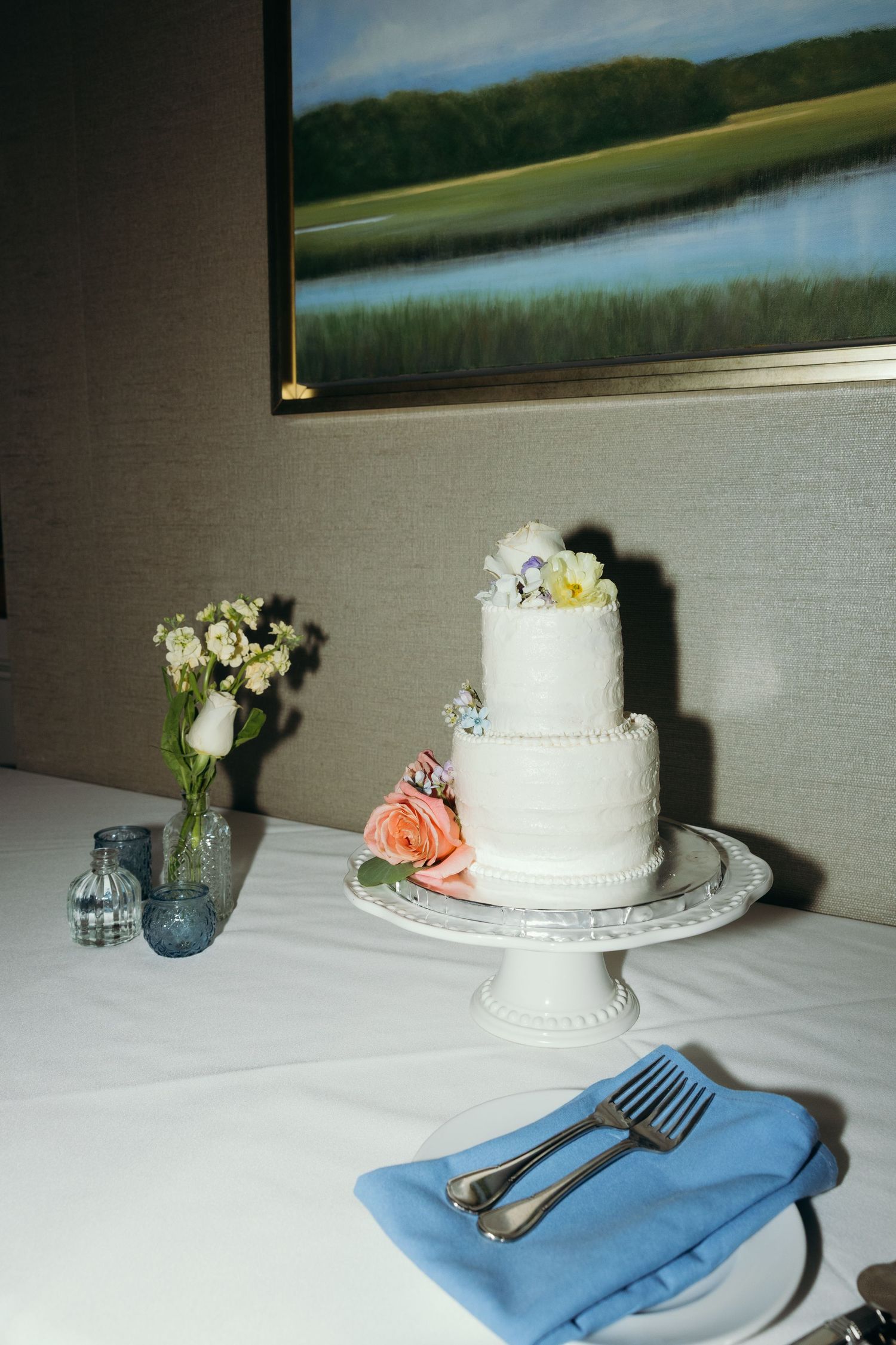 White tiered wedding cake with floral decorations displayed on a table with blue napkins.