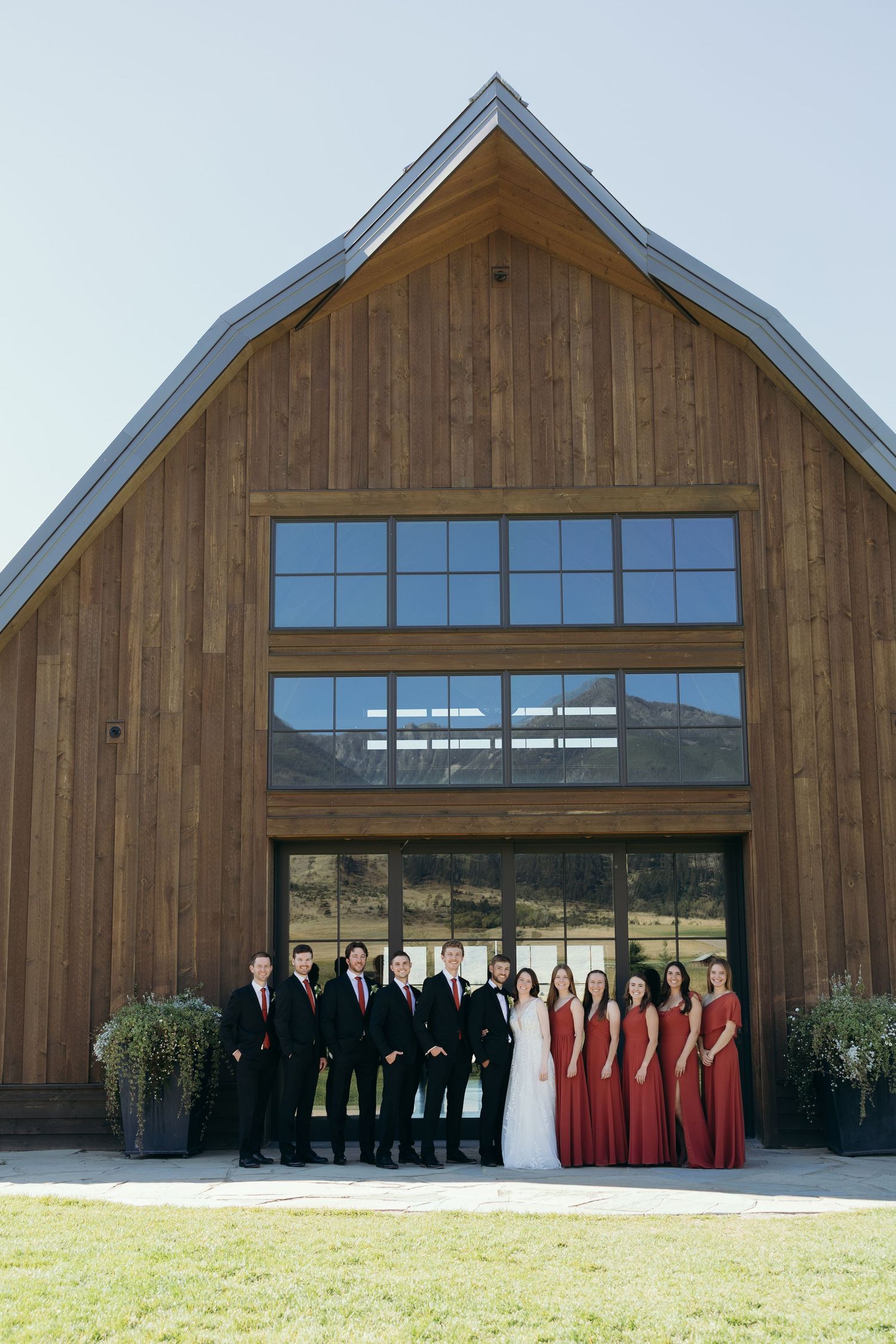 Wedding party members in red dresses and black suits stand against a modern barn exterior.