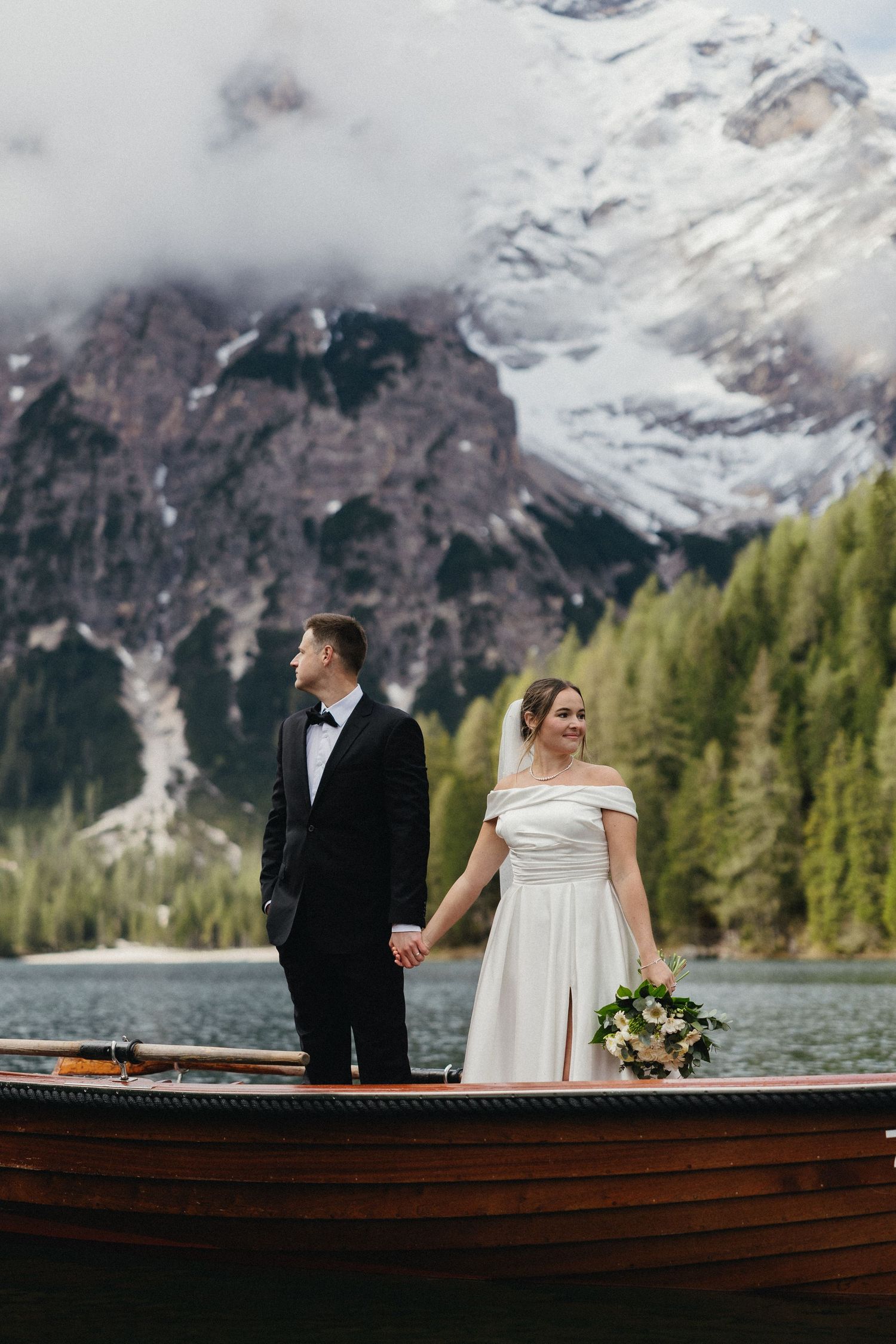 Couple in wedding attire standing on wooden boat with snow-capped mountains in background.