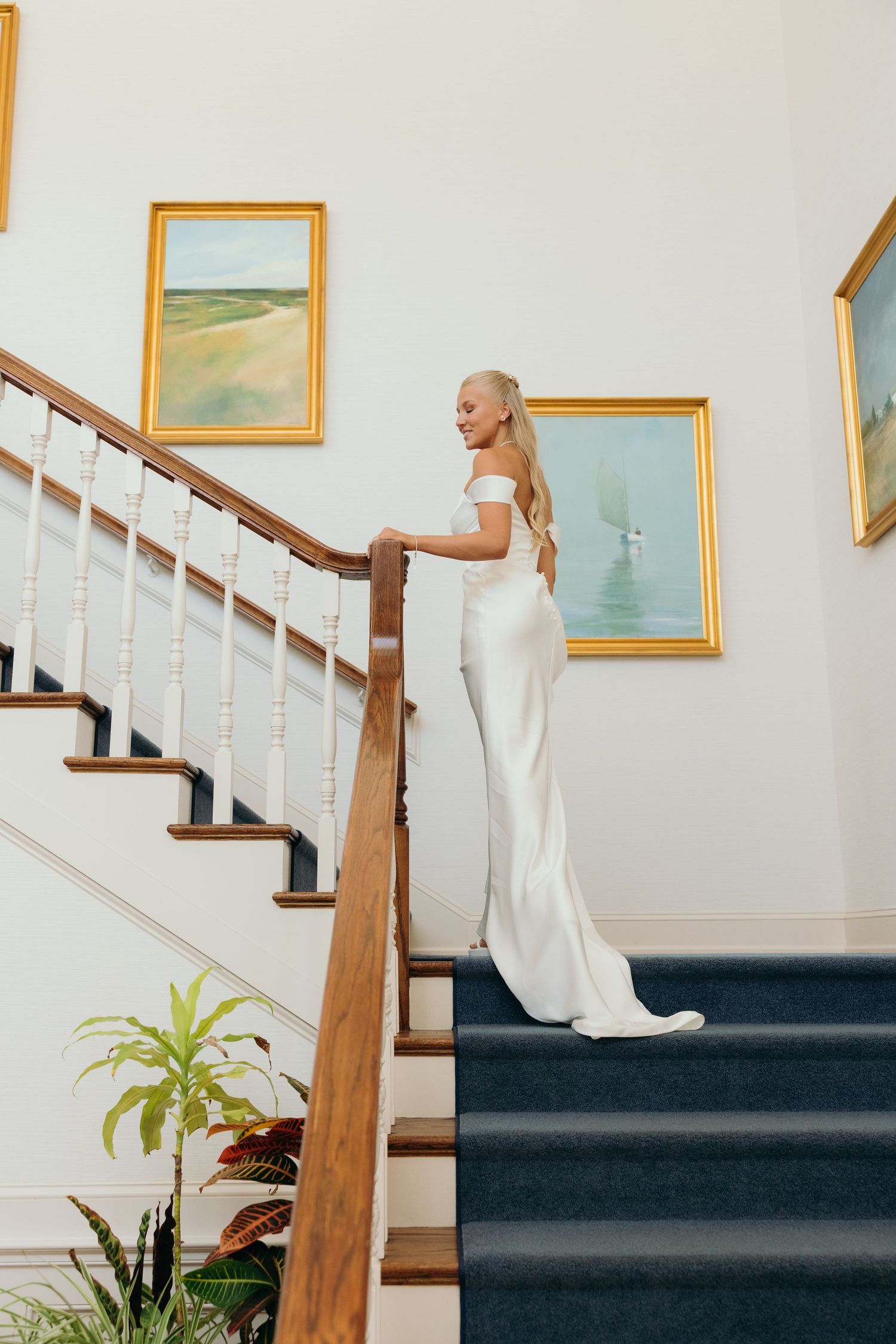 A bride in a white wedding gown poses on an elegant staircase with framed artwork.