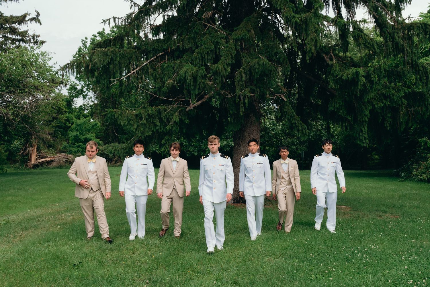 Wedding party in white and beige outfits standing on a grassy lawn with trees in the background.