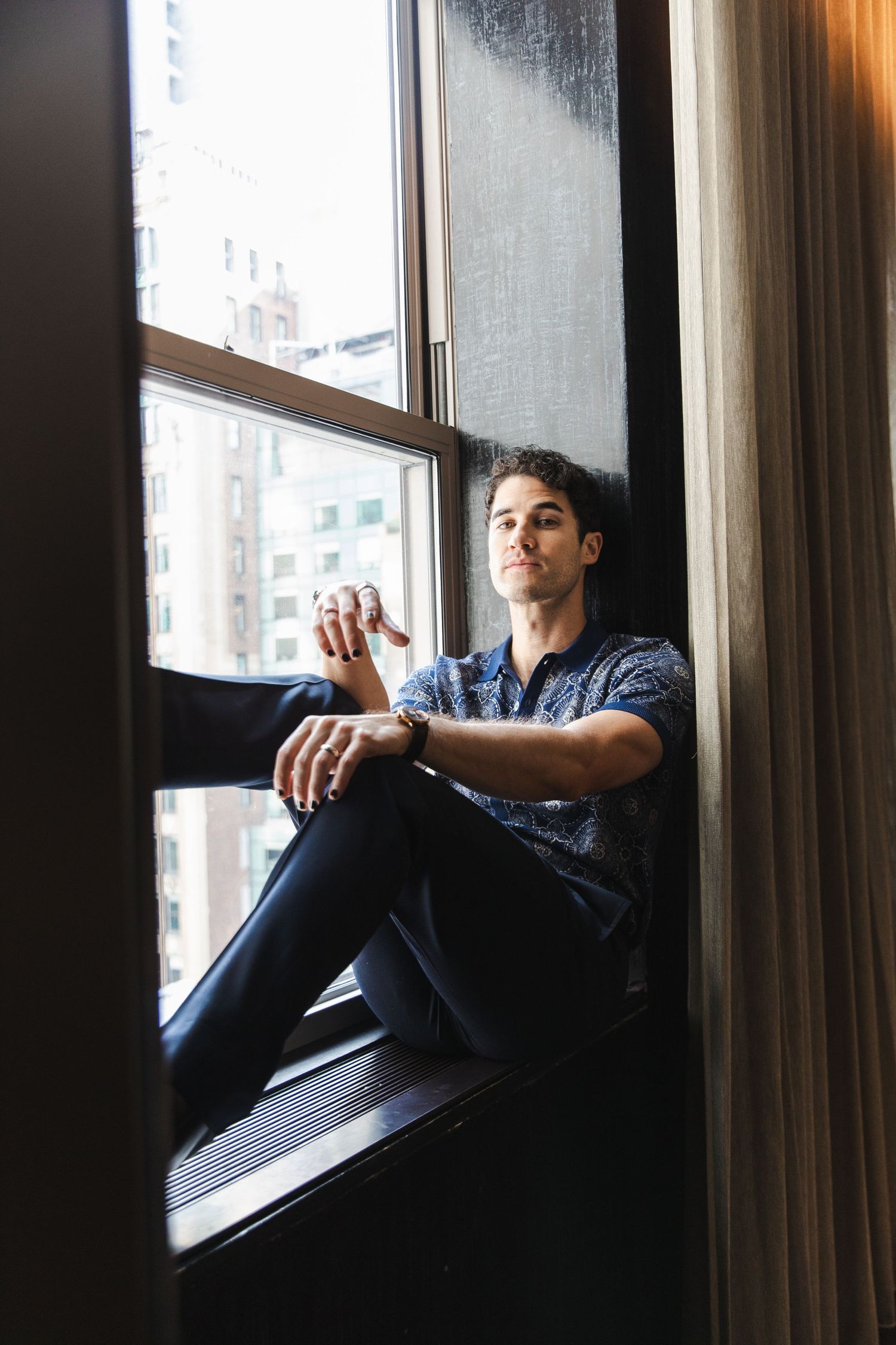Young man in floral shirt sitting on window sill, gazing thoughtfully with city buildings visible outside.