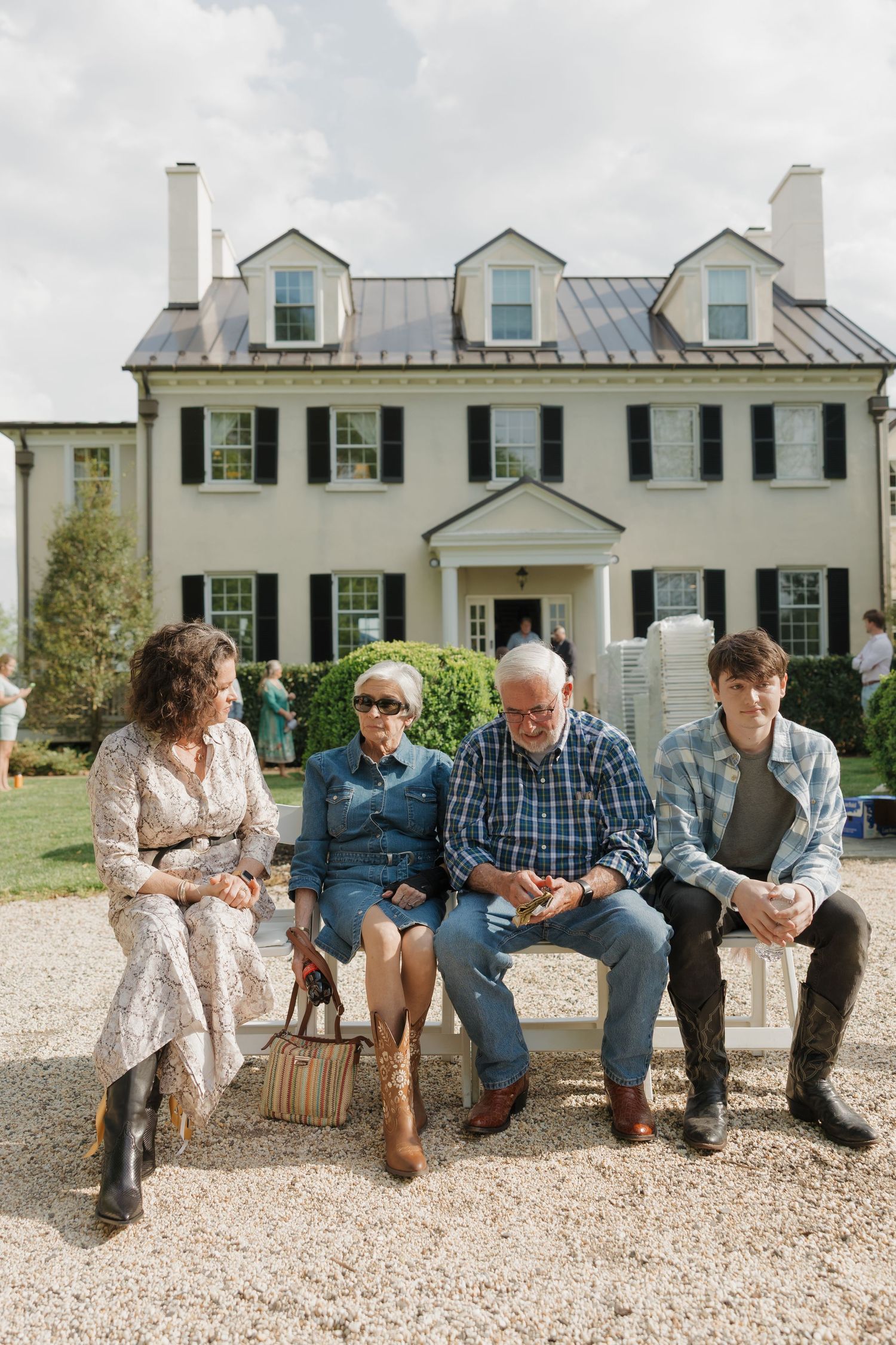 Four people sitting on a bench outside a large white colonial-style home during a sunny day.