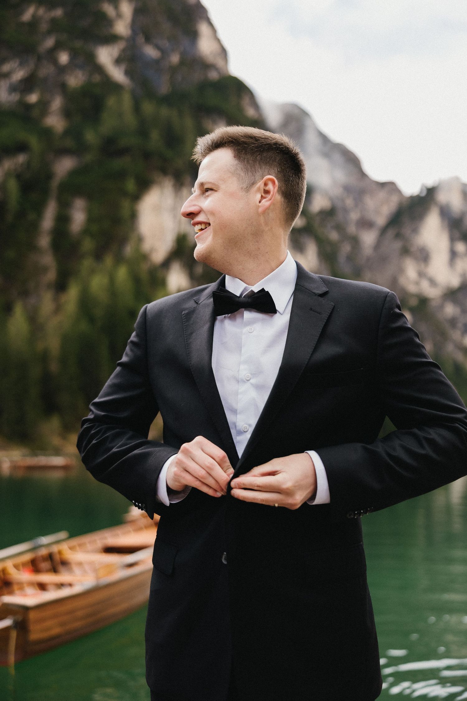 Groom in classic black tuxedo smiling by mountain lake with wooden boats in background.