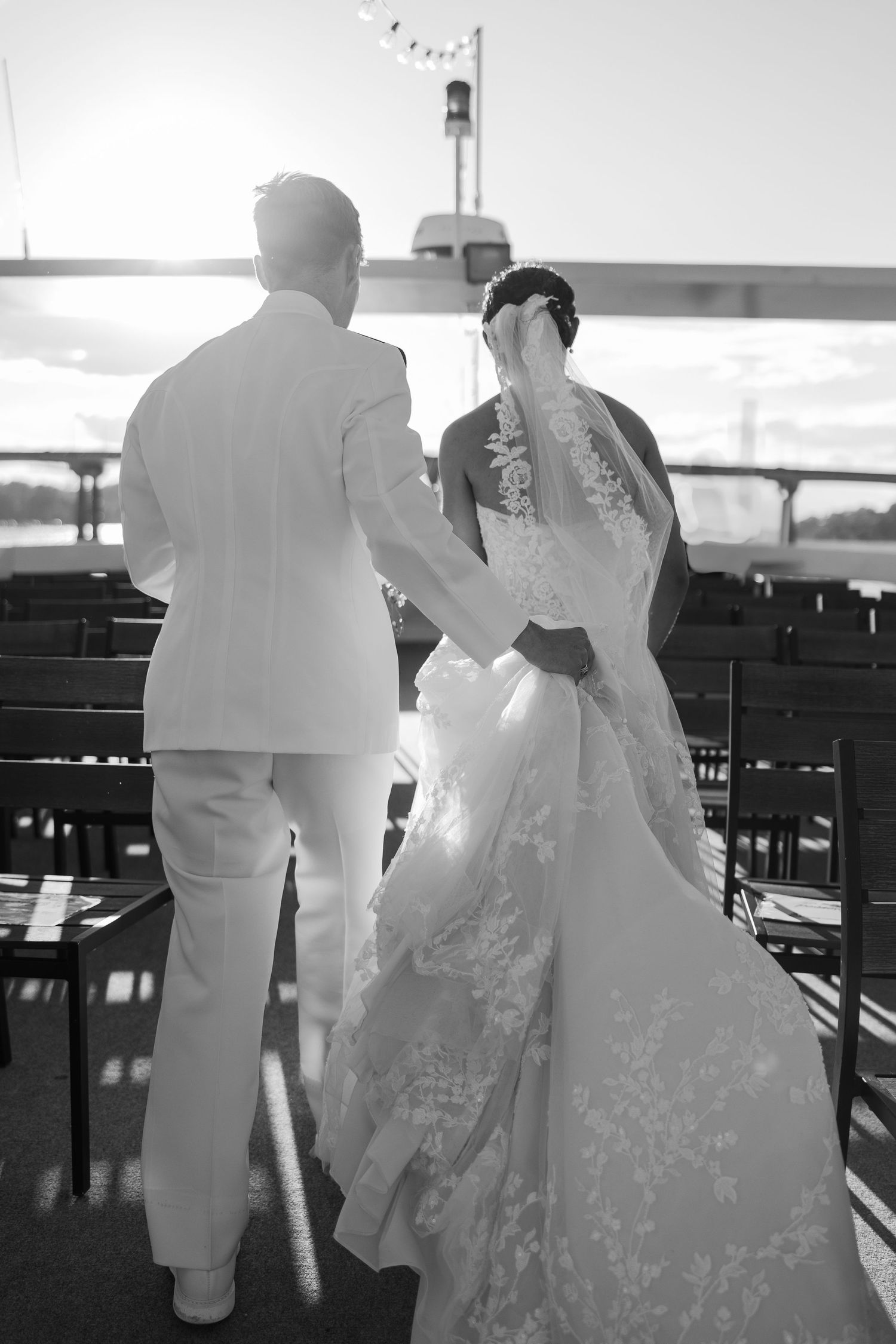 A couple in white formal attire walks along a deck with railing at sunset.