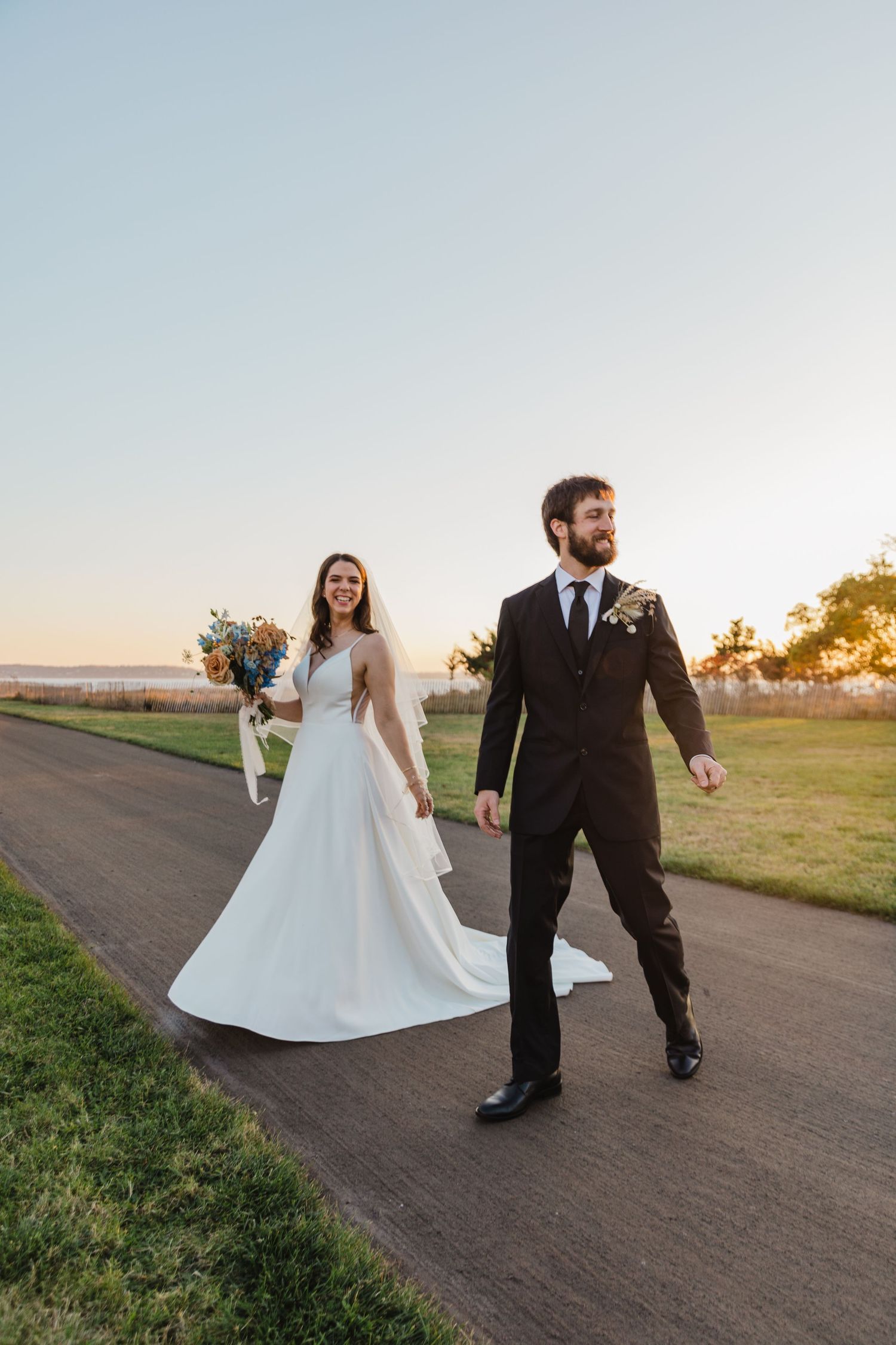 Newlyweds walking together on a path at sunset, bride holding a bouquet.