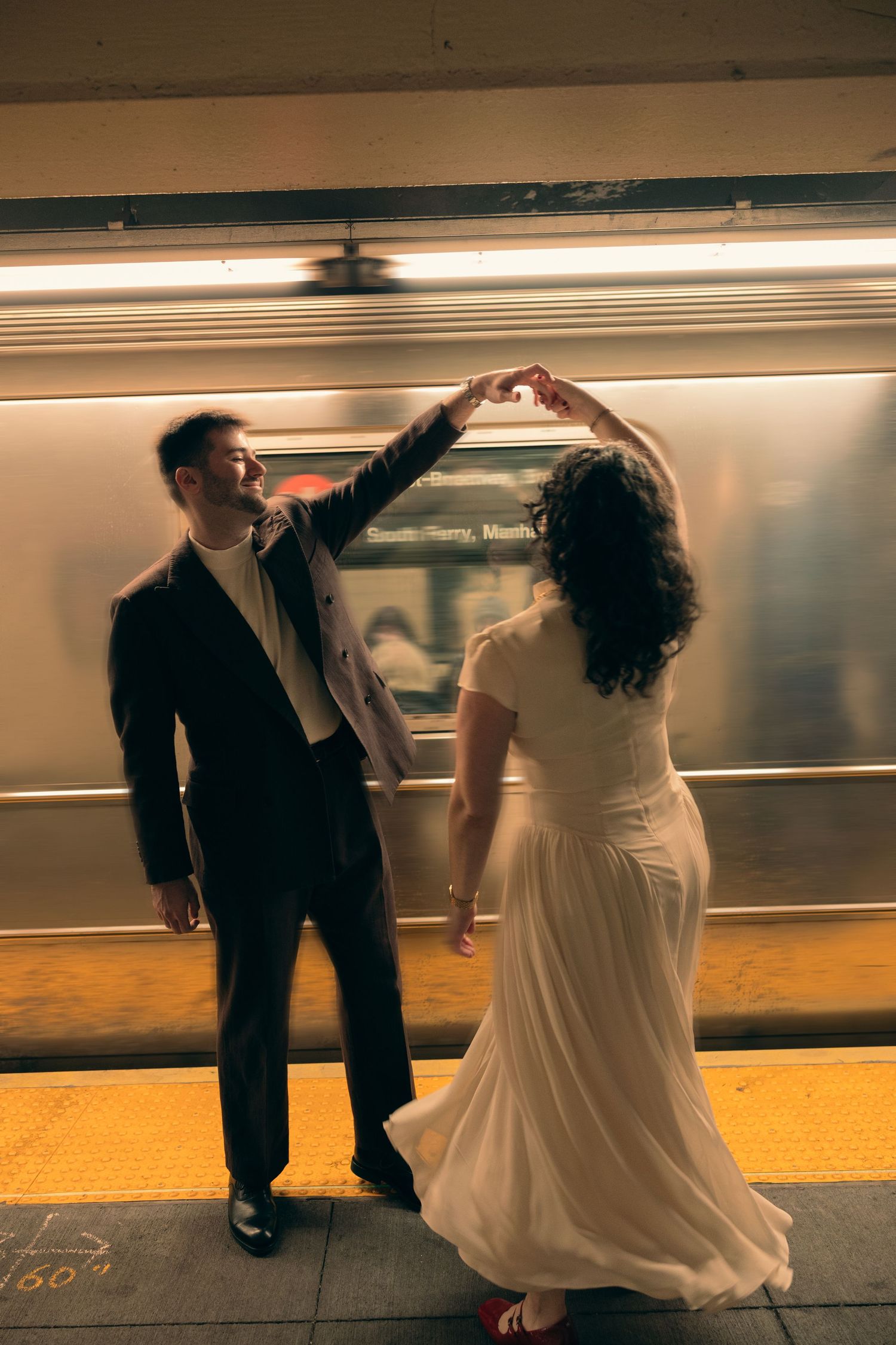 A couple in wedding attire dances on a subway platform as a train blurs past in warm lighting.
