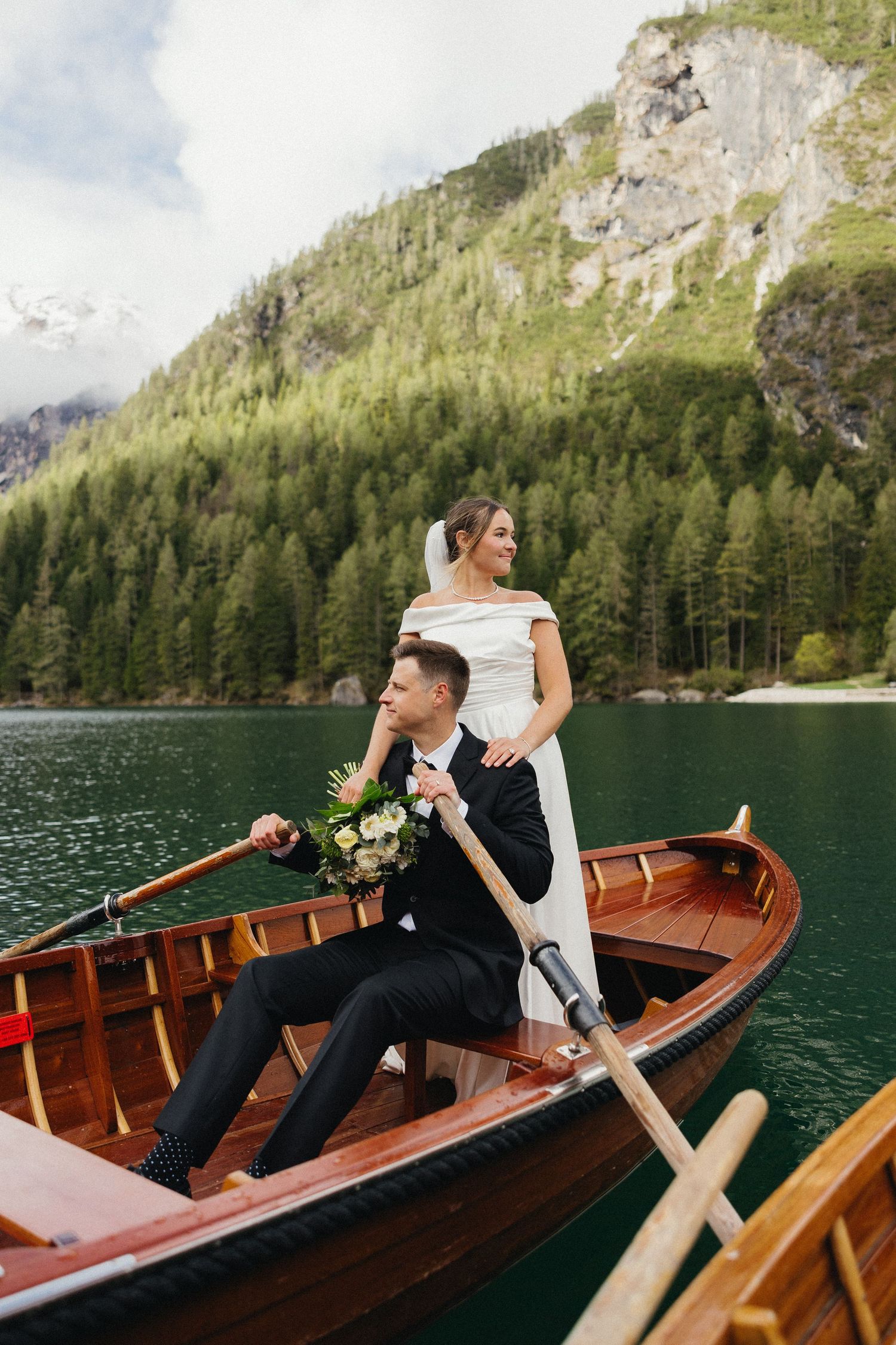 A couple enjoys a romantic boat ride on a serene mountain lake in Italy surrounded by evergreen forests and rocky cliffs.