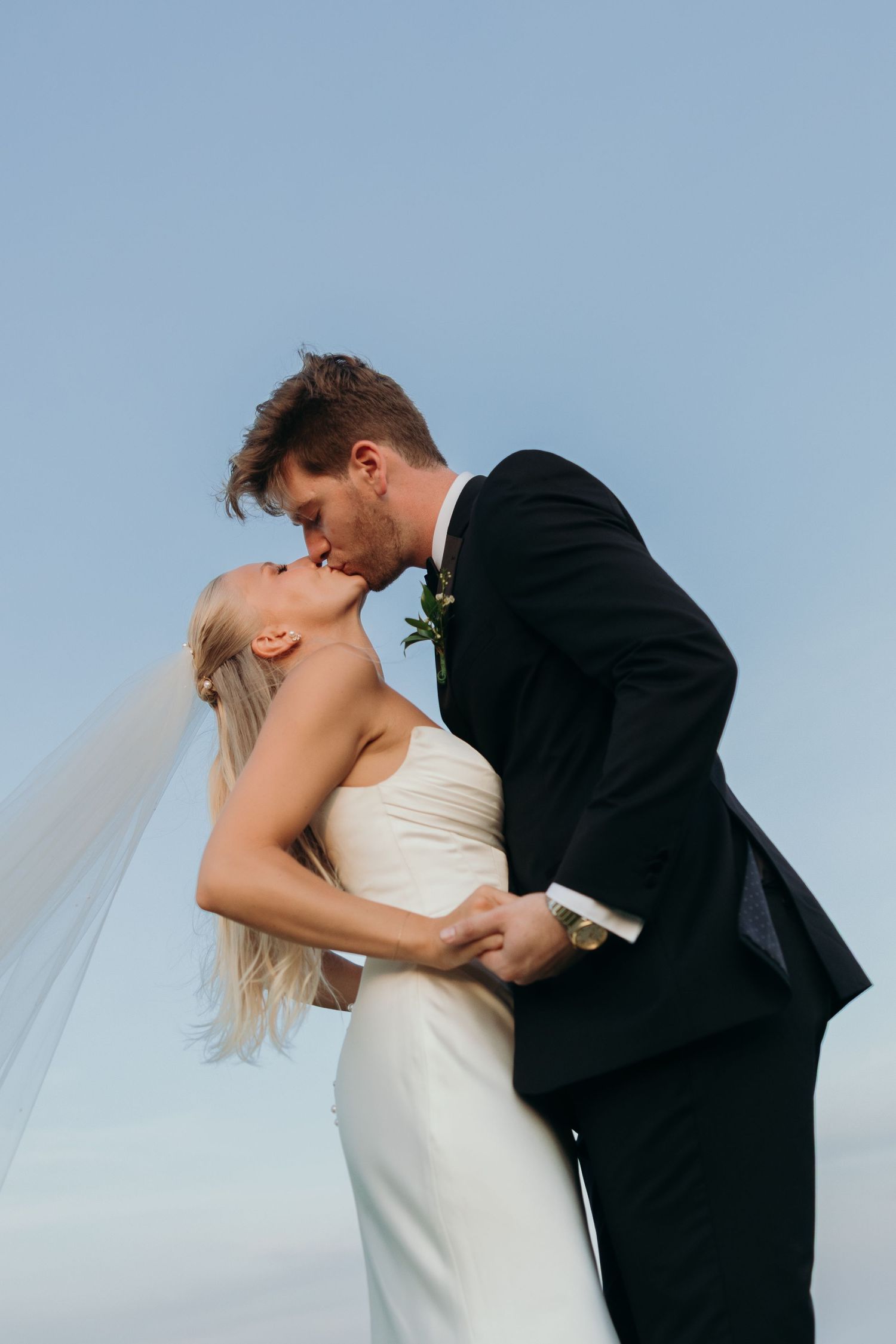 Romantic wedding couple embracing against a blue sky backdrop.
